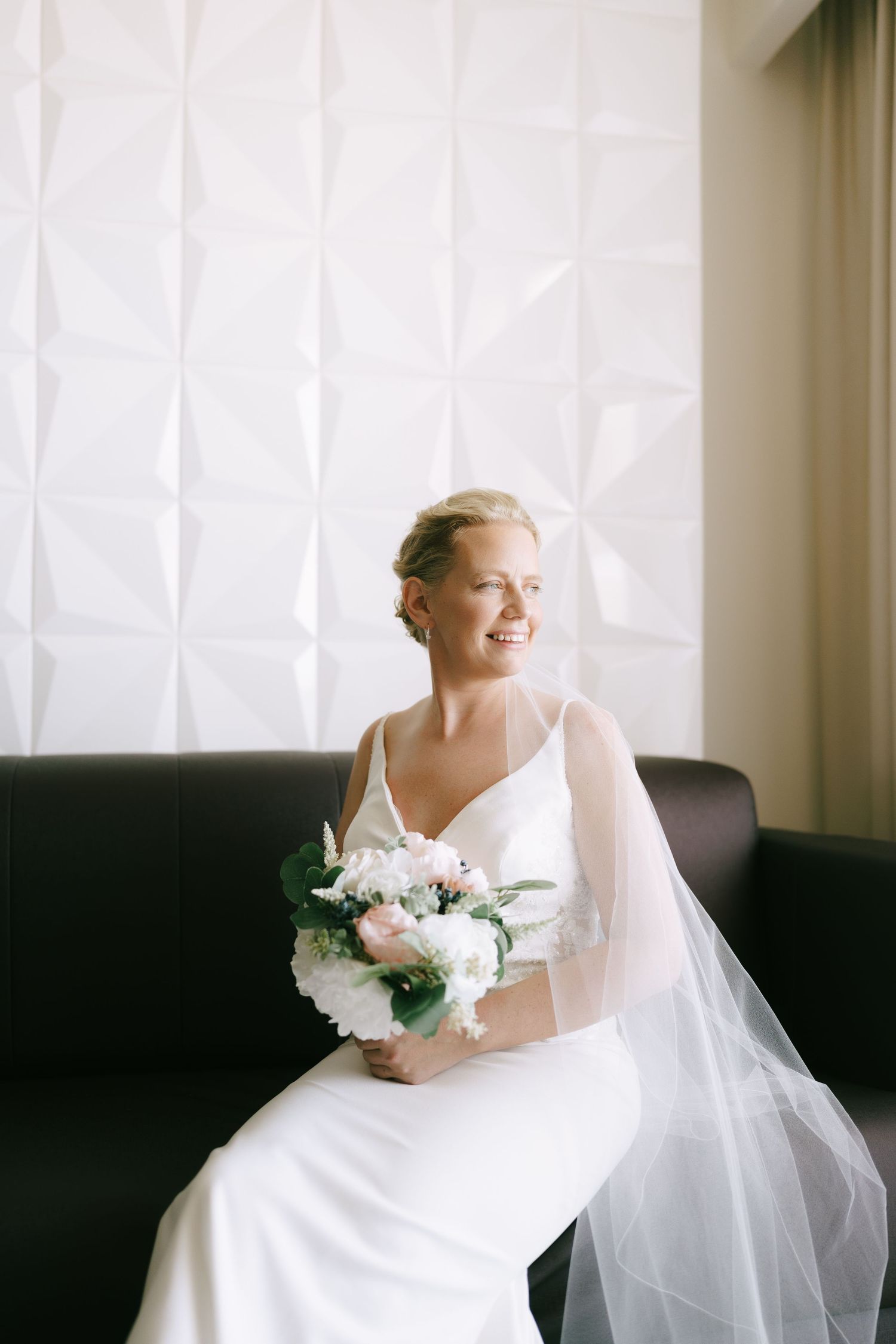 Bride in white dress holding bouquet by window in bright artistic wedding photo series.