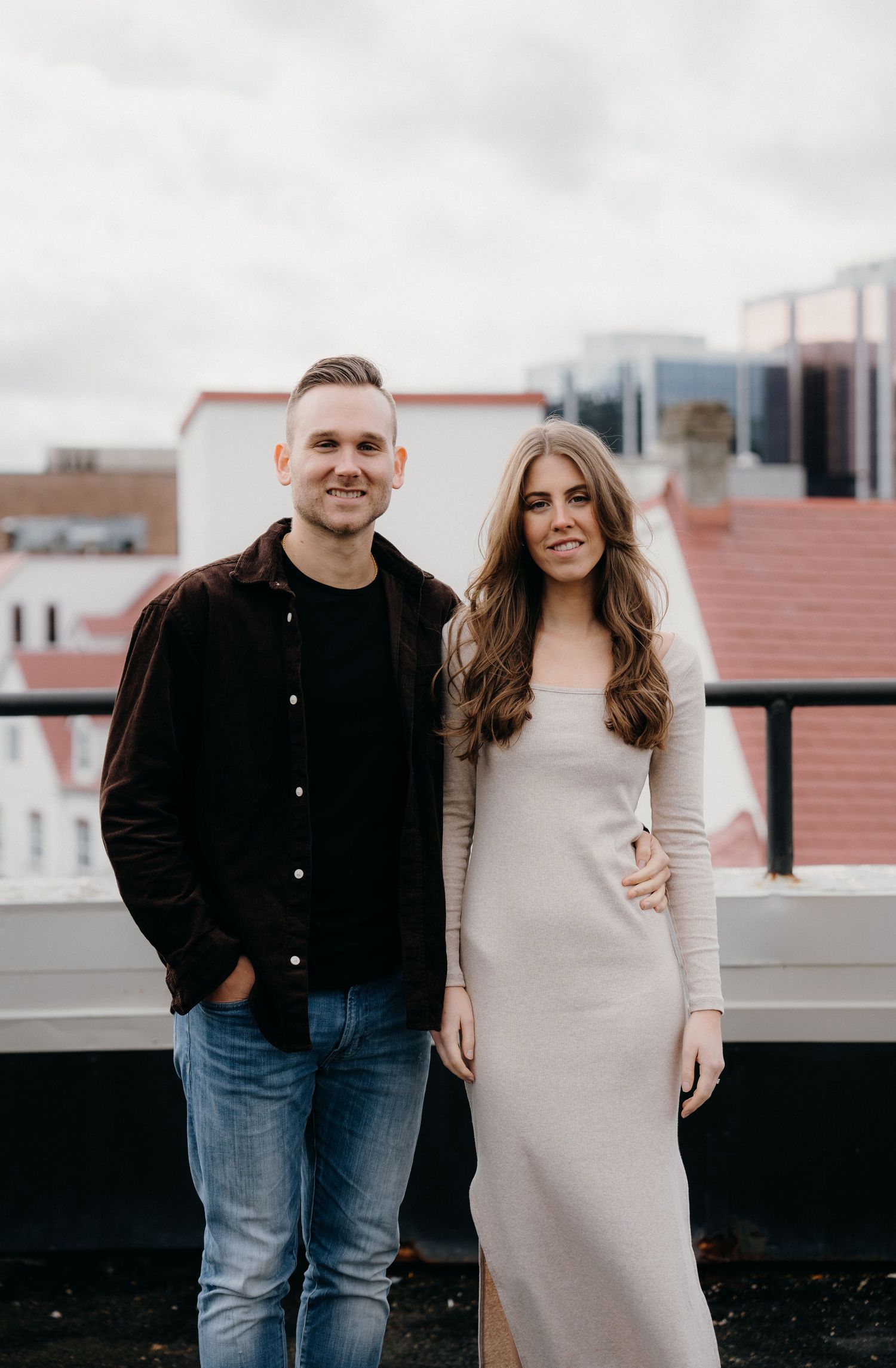 Couple poses together on urban rooftop, wearing casual clothes against cloudy sky.