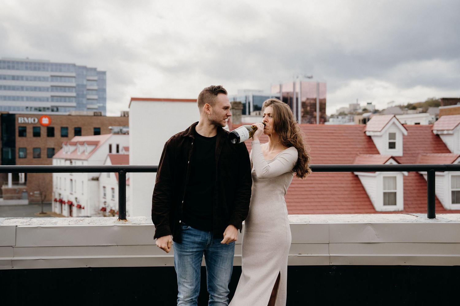 Two people stand together on a rooftop with an urban landscape behind them.