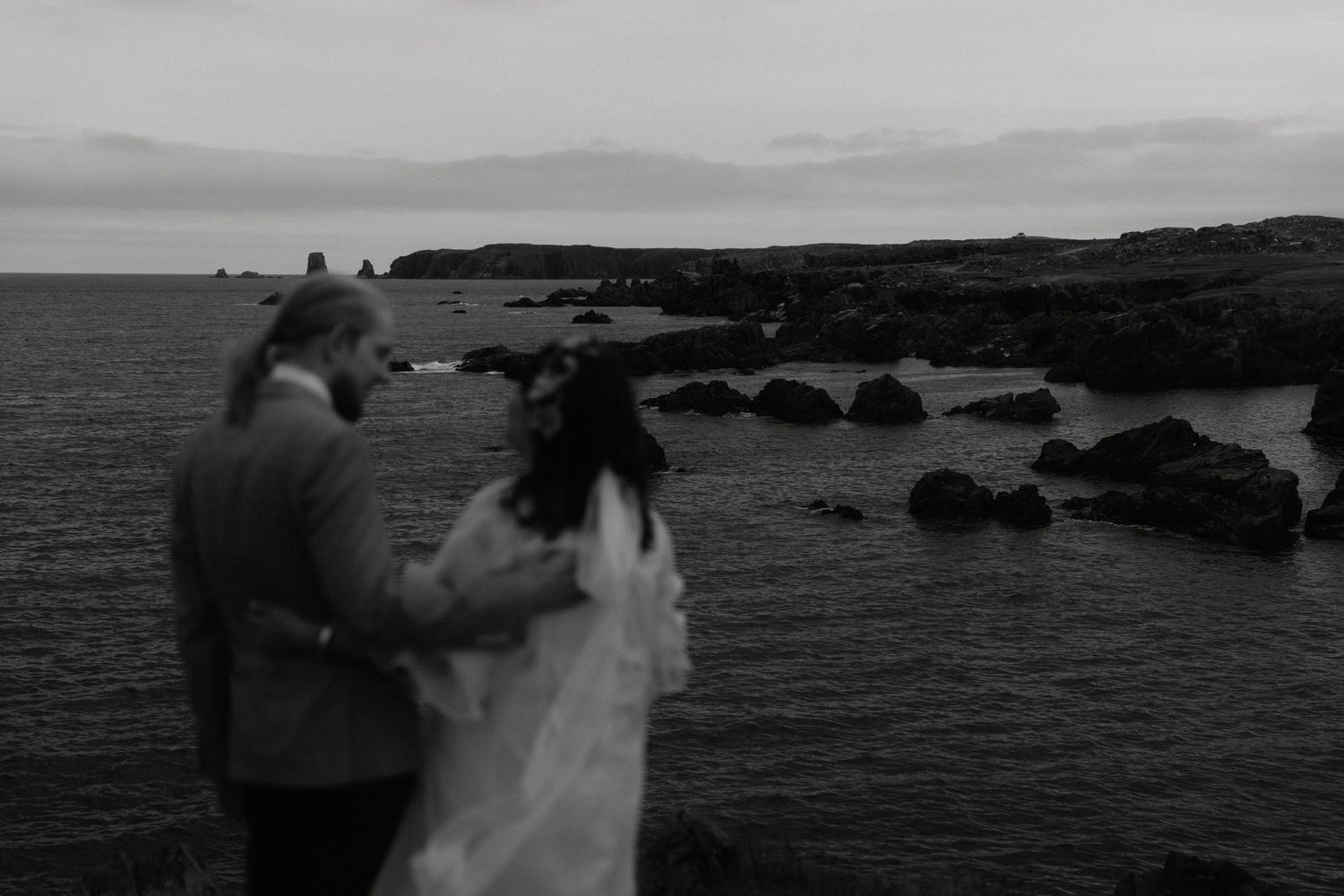 A couple stands in silhouette against a rocky coastline and ocean view in black and white.