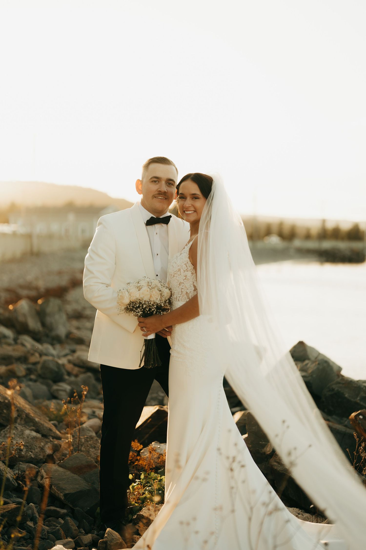 Newlyweds share an intimate moment on a rocky shoreline during golden hour in elegant wedding attire.