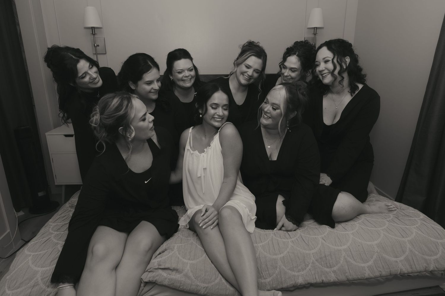 A black and white photo of a group of women in dark clothing laughing together while sitting on a bed.
