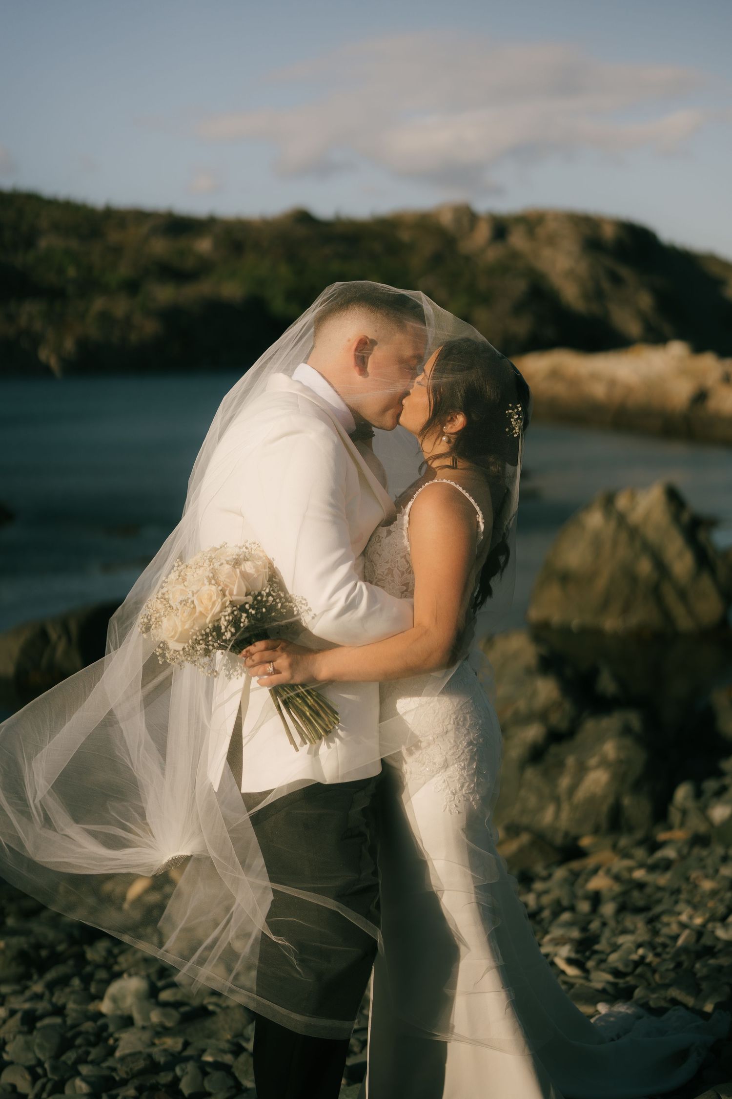 A romantic newlywed couple embraces on a rocky beach at sunset with the bride's veil flowing in the wind.