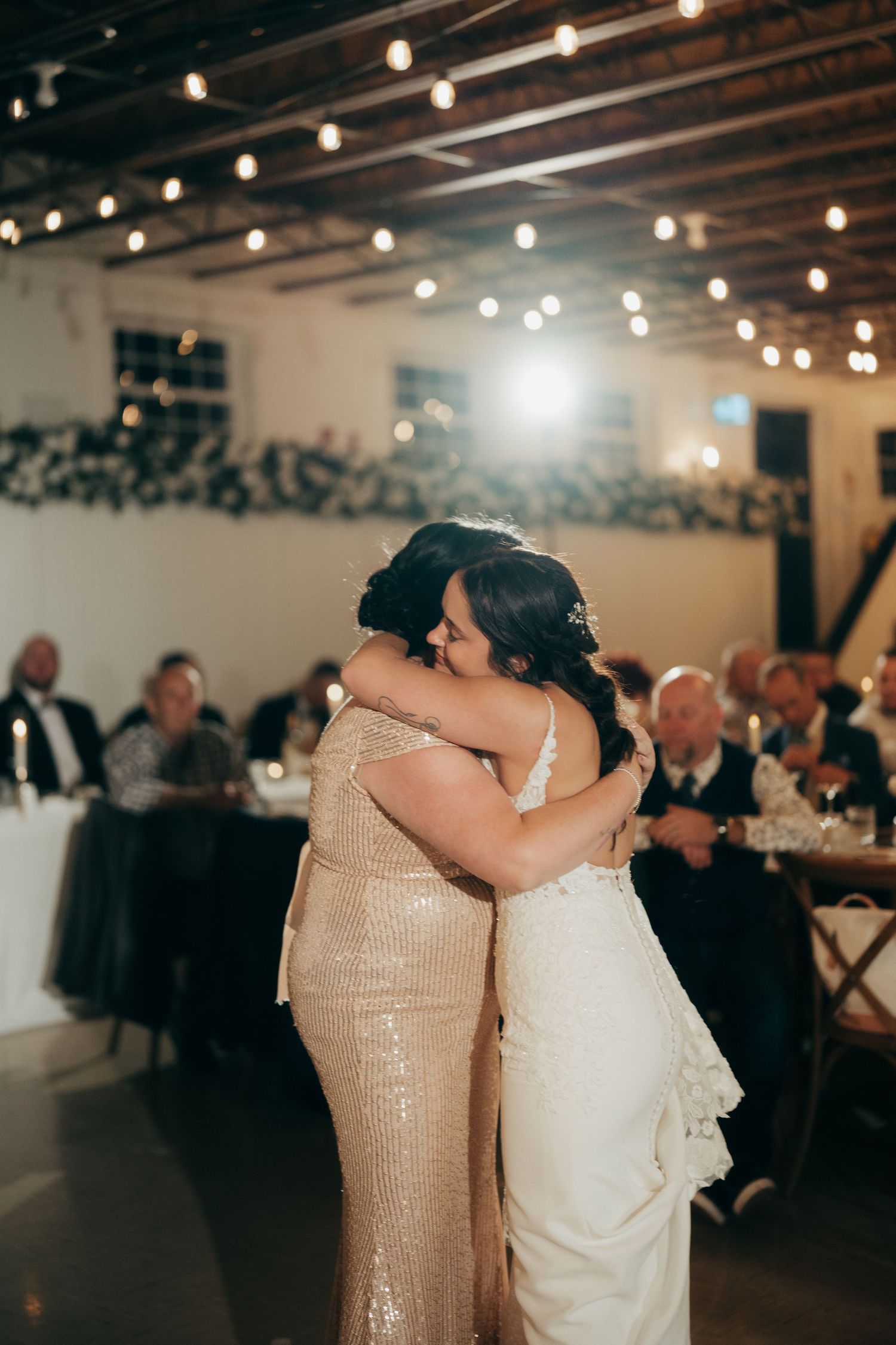 Mother and bride share an emotional embrace on the dance floor during wedding reception under warm string lights.