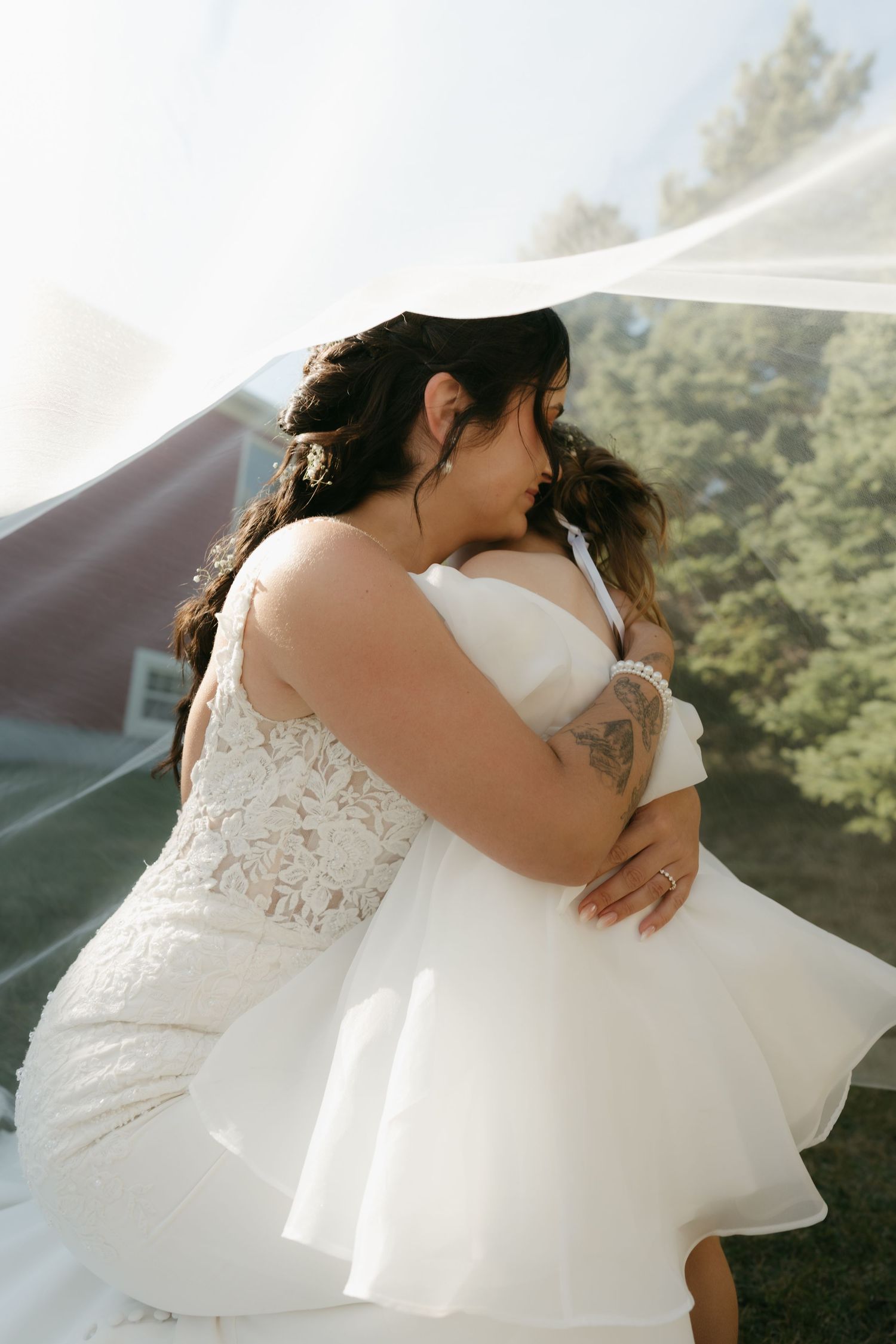 Two people in white wedding attire embrace lovingly in a sun-drenched outdoor setting with trees in the background.