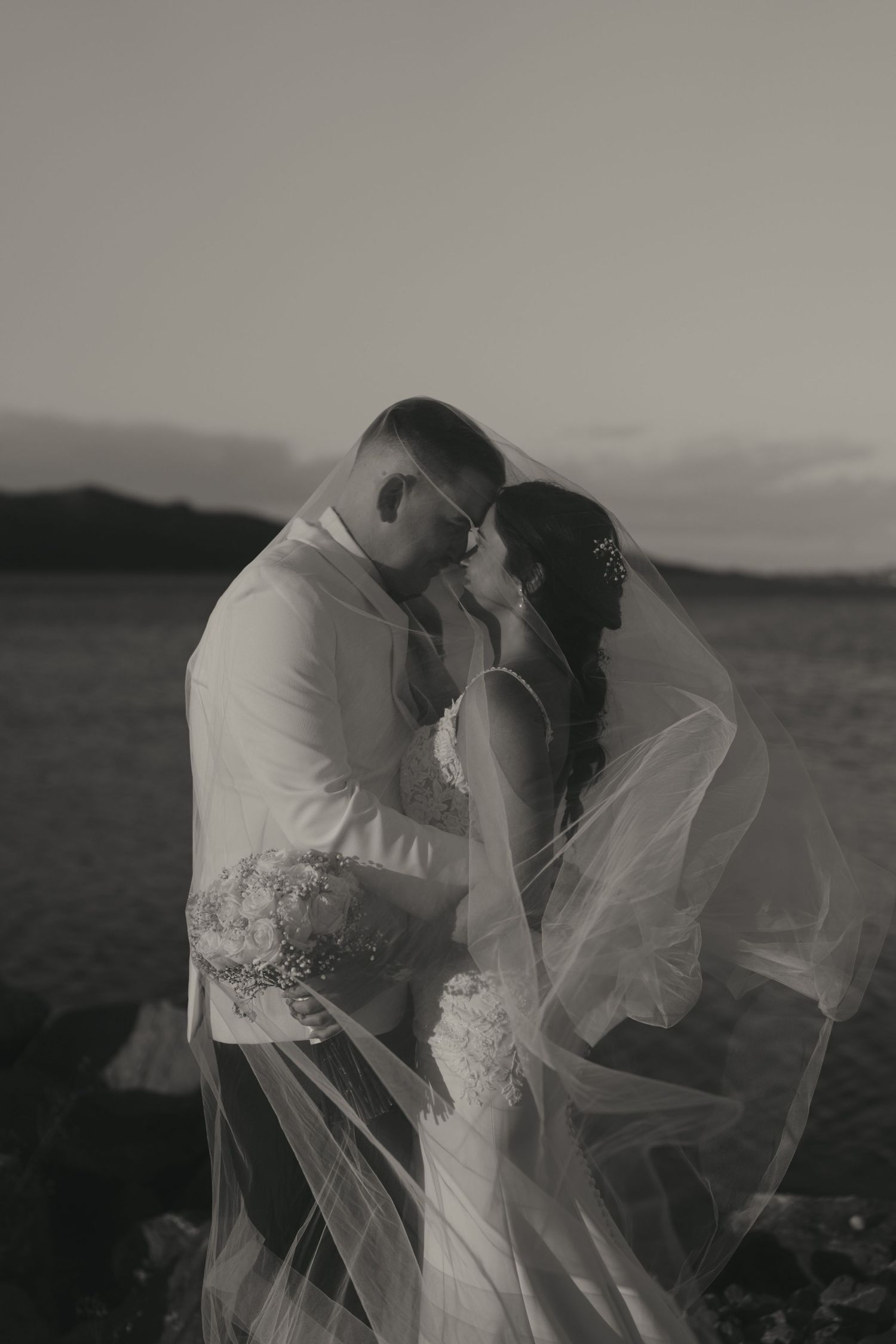 An intimate black and white wedding portrait captures a tender moment between newlyweds as a veil flows in the breeze.