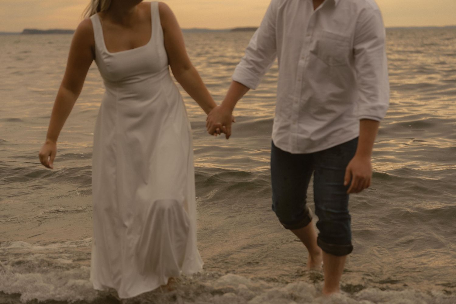 A couple holds hands while walking in shallow ocean water at sunset, with the woman wearing a white dress.