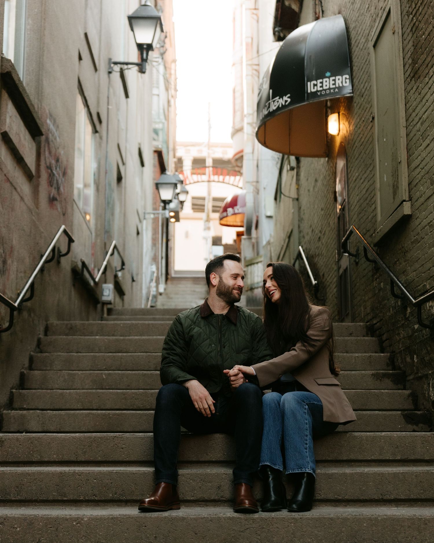 Couple sitting on urban alley steps, smiling at each other, with street lamps and an Iceberg umbrella overhead.