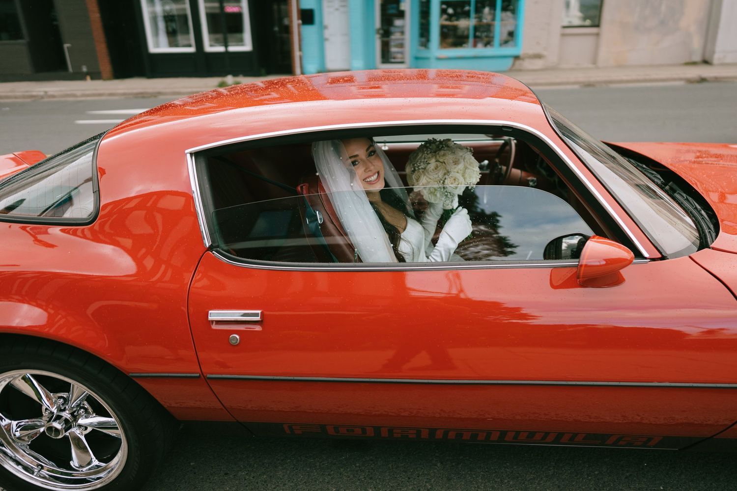 Bride in wedding dress and veil sitting in a classic red muscle car on a city street.