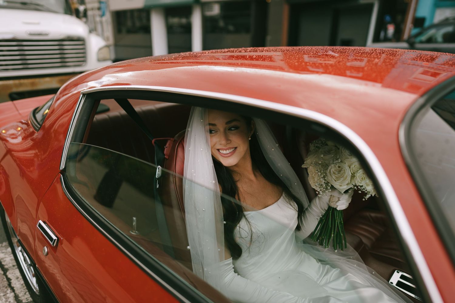 Smiling bride in white gown and veil holding bouquet inside a classic red car on her wedding day.