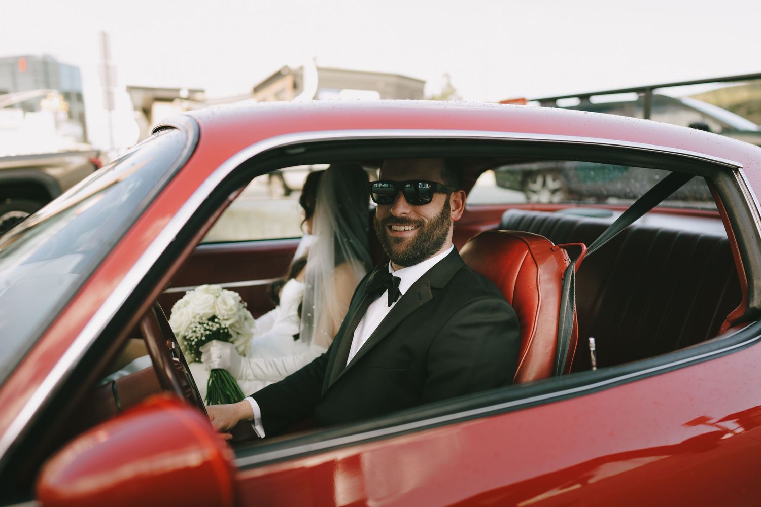 Smiling groom in black tuxedo and sunglasses sitting with bride holding bouquet in a classic red car.