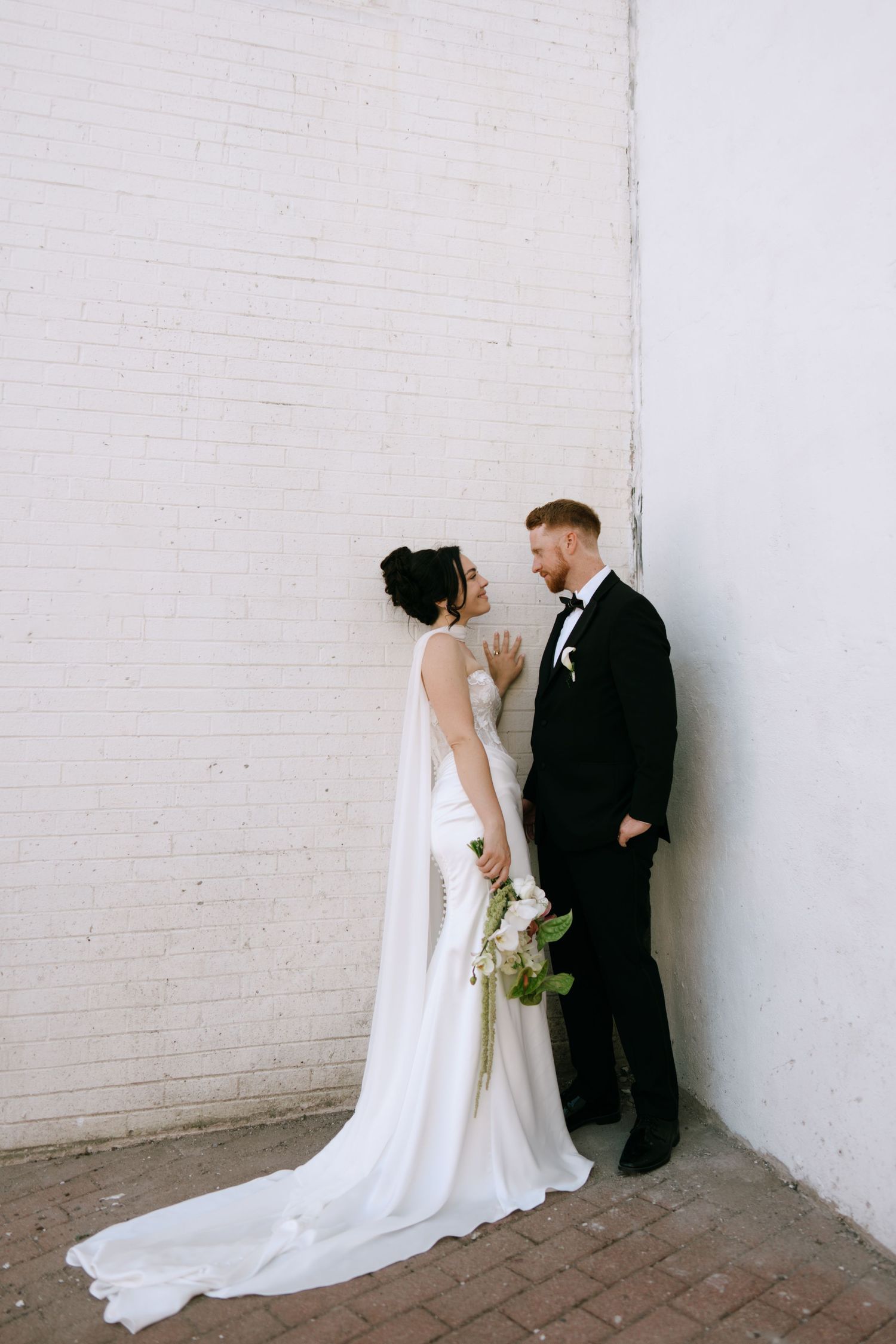 Bride and groom in elegant wedding attire pose against a white brick wall, sharing an intimate moment.
