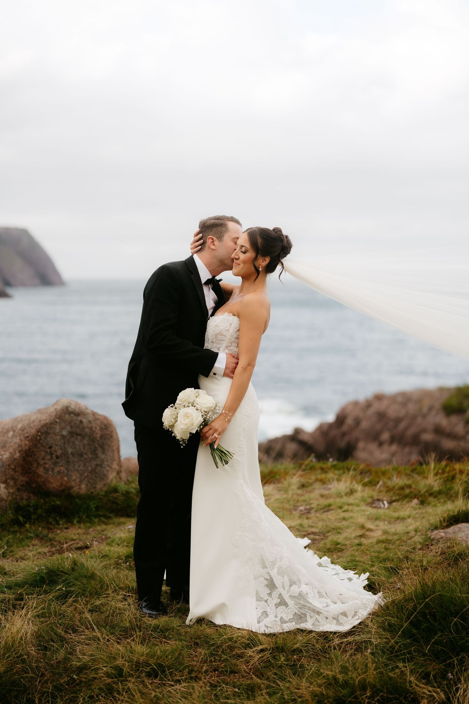 Bride and groom share a romantic kiss on a scenic coastal cliff, with veil flowing in the wind and ocean backdrop.