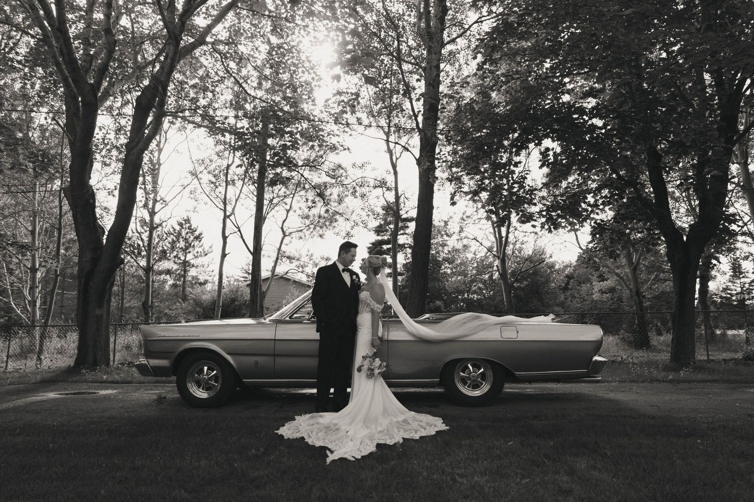 Black and white photo of bride and groom standing beside a classic convertible car surrounded by trees.