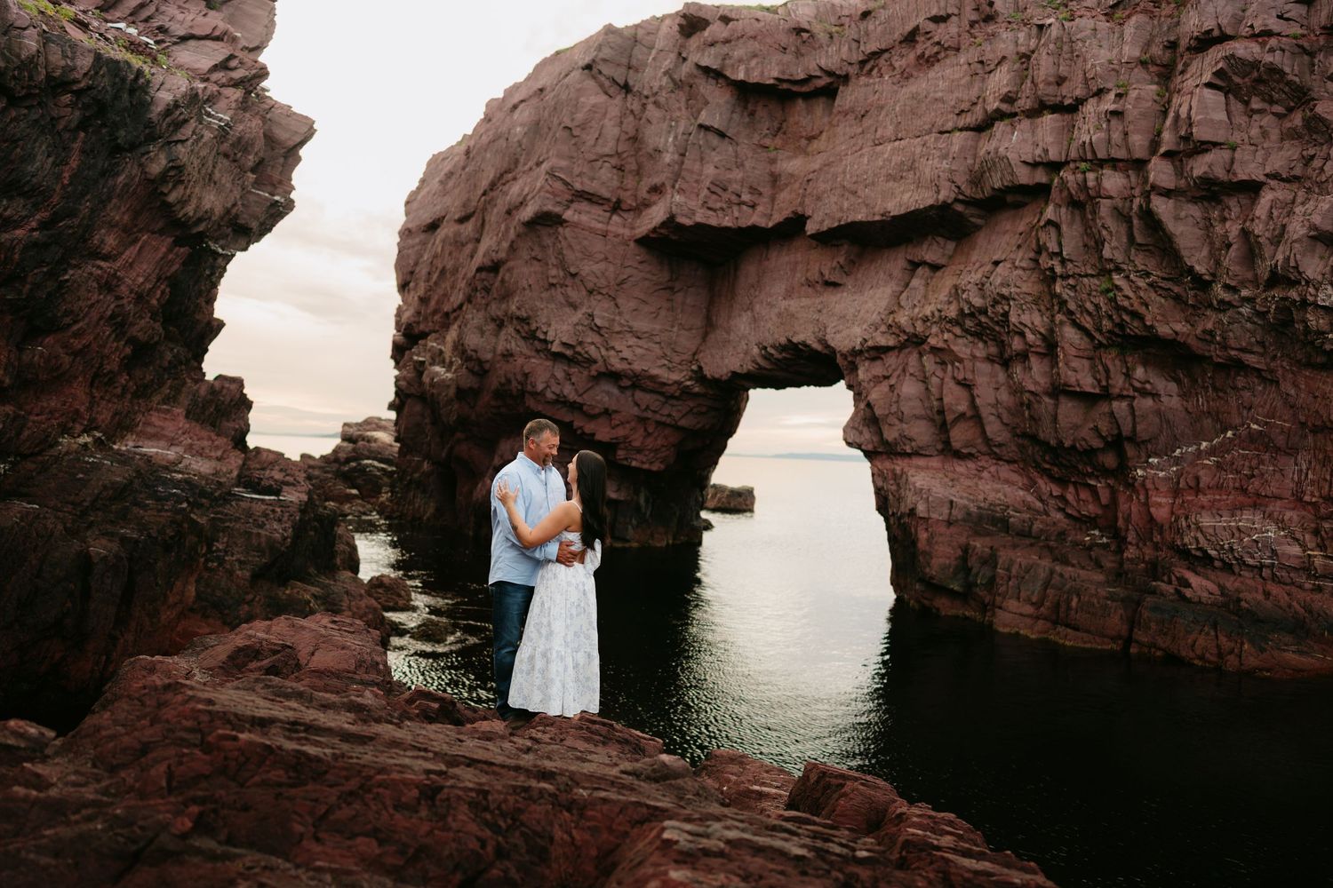 Couple embracing under a natural red rock arch by the ocean at sunset, romantic coastal engagement photo.