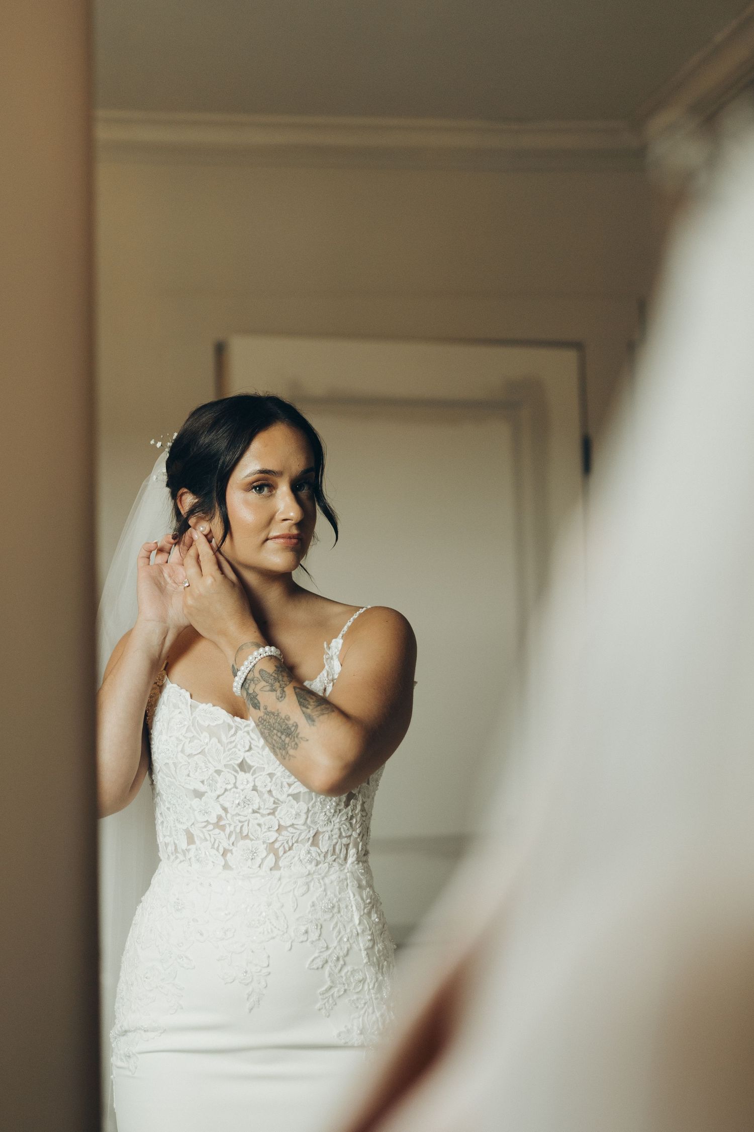 A bride in a white lace wedding dress puts on earrings while getting ready in a softly lit room.