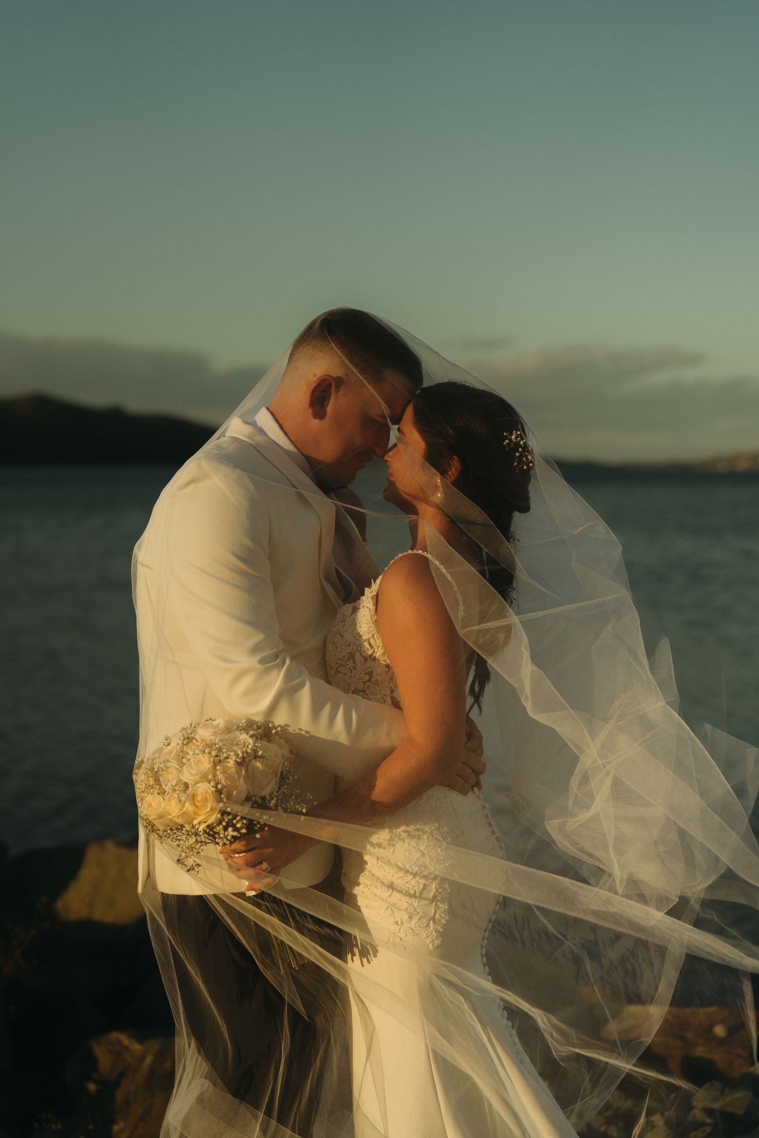 A couple shares a romantic kiss at sunset by the ocean while a white veil flows in the breeze.