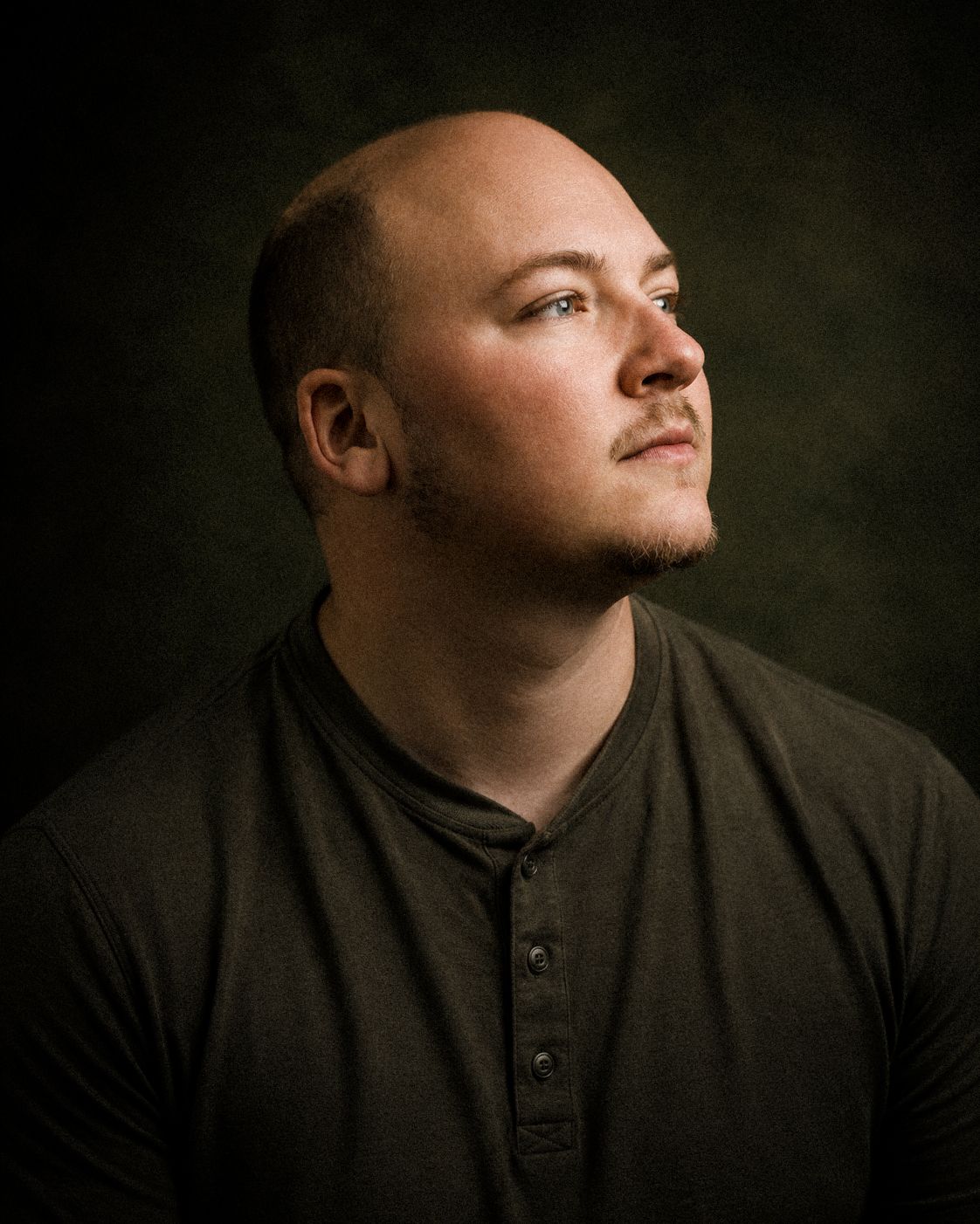 A bald man with a goatee wearing a dark henley shirt gazes upward in a dramatic studio portrait.