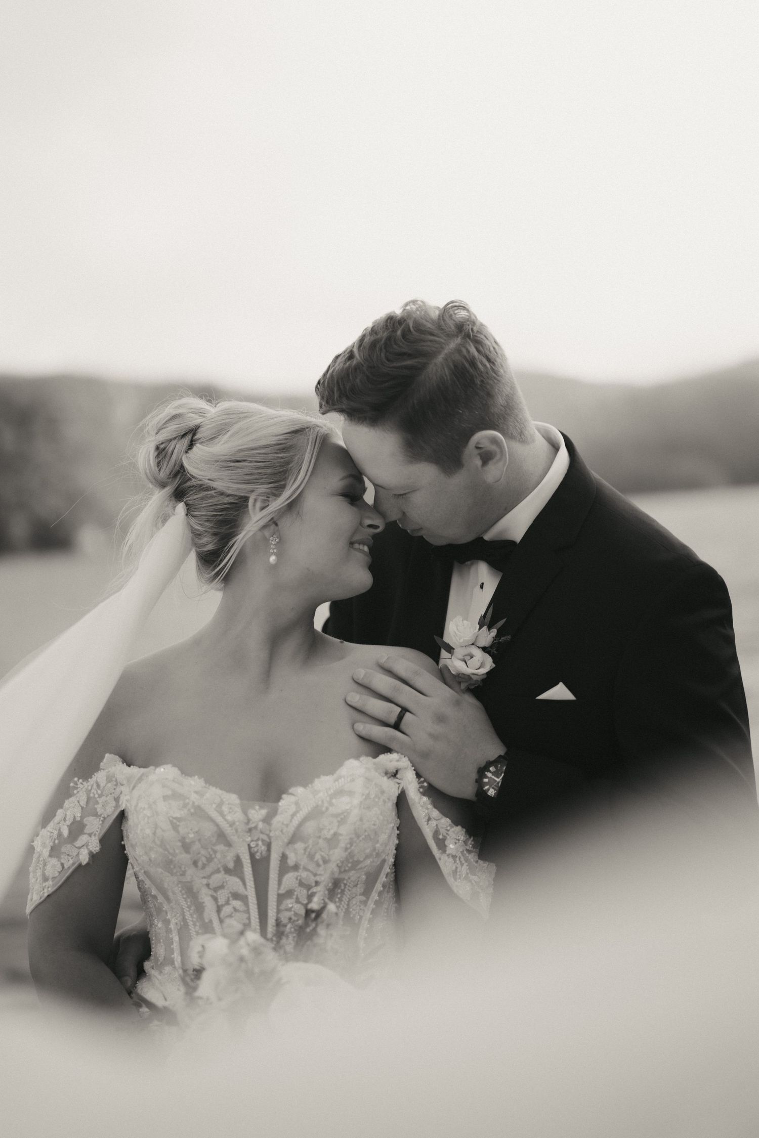 Black and white photo of a bride and groom sharing an intimate moment, groom in tuxedo kissing bride in lace gown.