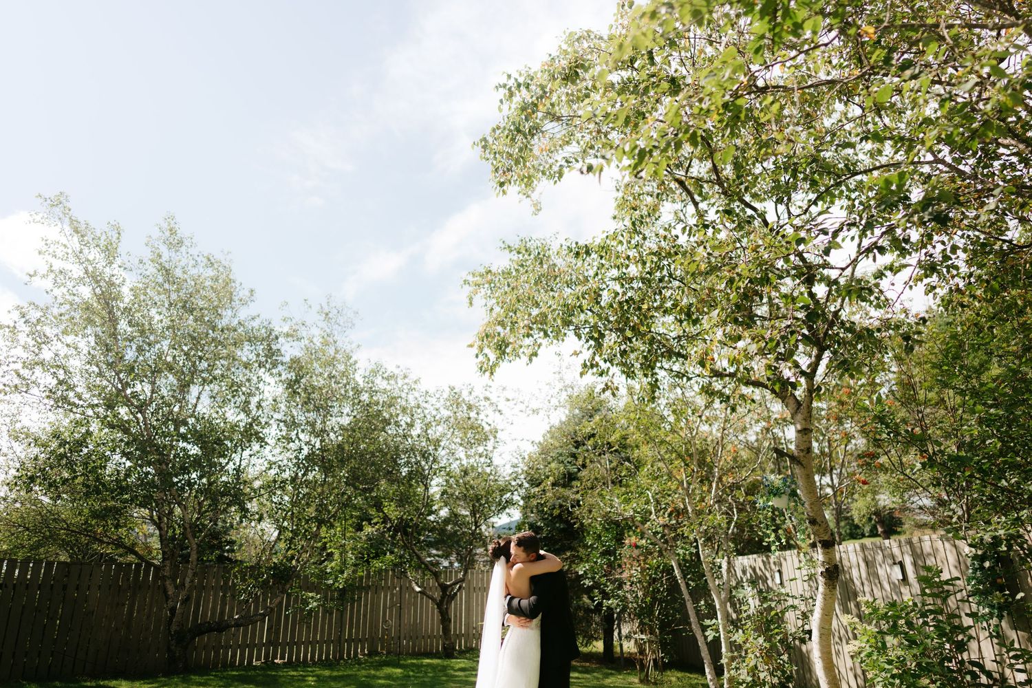 A bride and groom embrace in a lush green backyard garden on their wedding day under a partly cloudy sky.