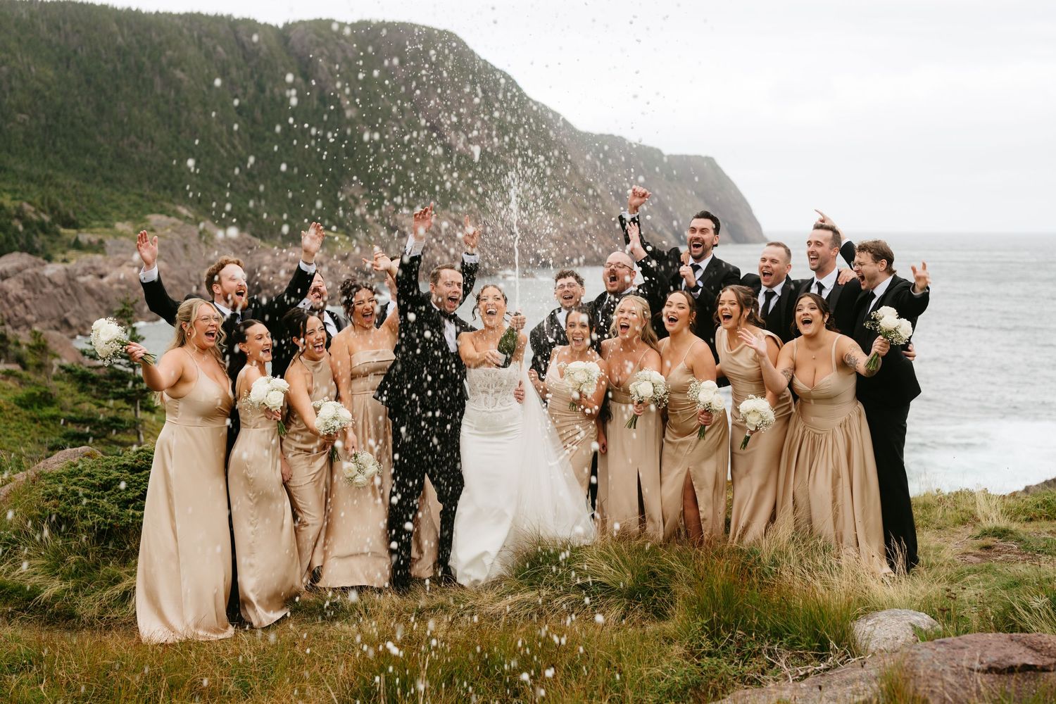 Wedding party celebrates outdoors near coastal cliffs, bridesmaids in gold dresses tossing confetti with ocean backdrop.