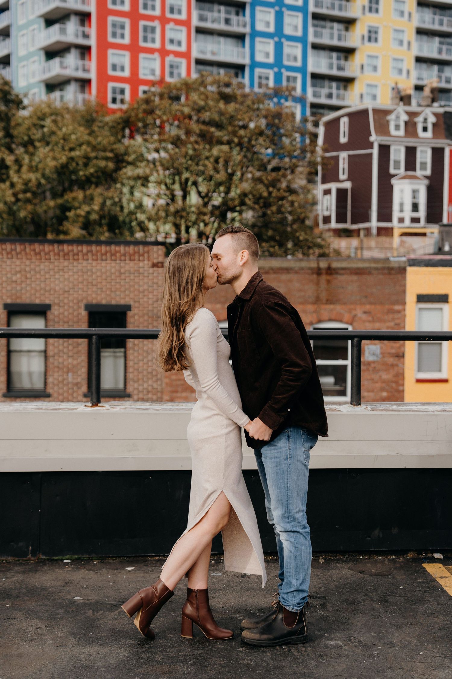 Intimate moment captured between couple on rooftop with city architecture and fall foliage in background.