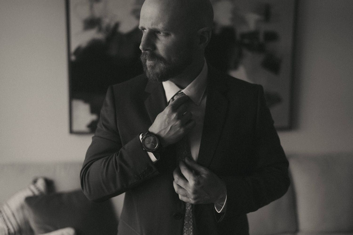 Groom adjusting tie and suit in dimly lit room before wedding ceremony.