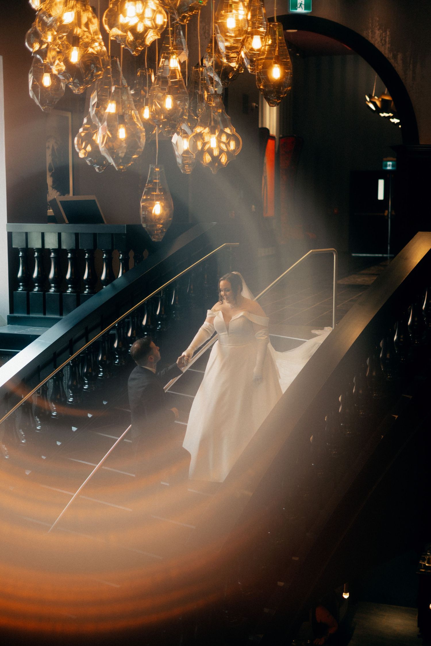 Artistic wedding photography shows silhouettes on a dark staircase illuminated by warm glowing chandeliers above.