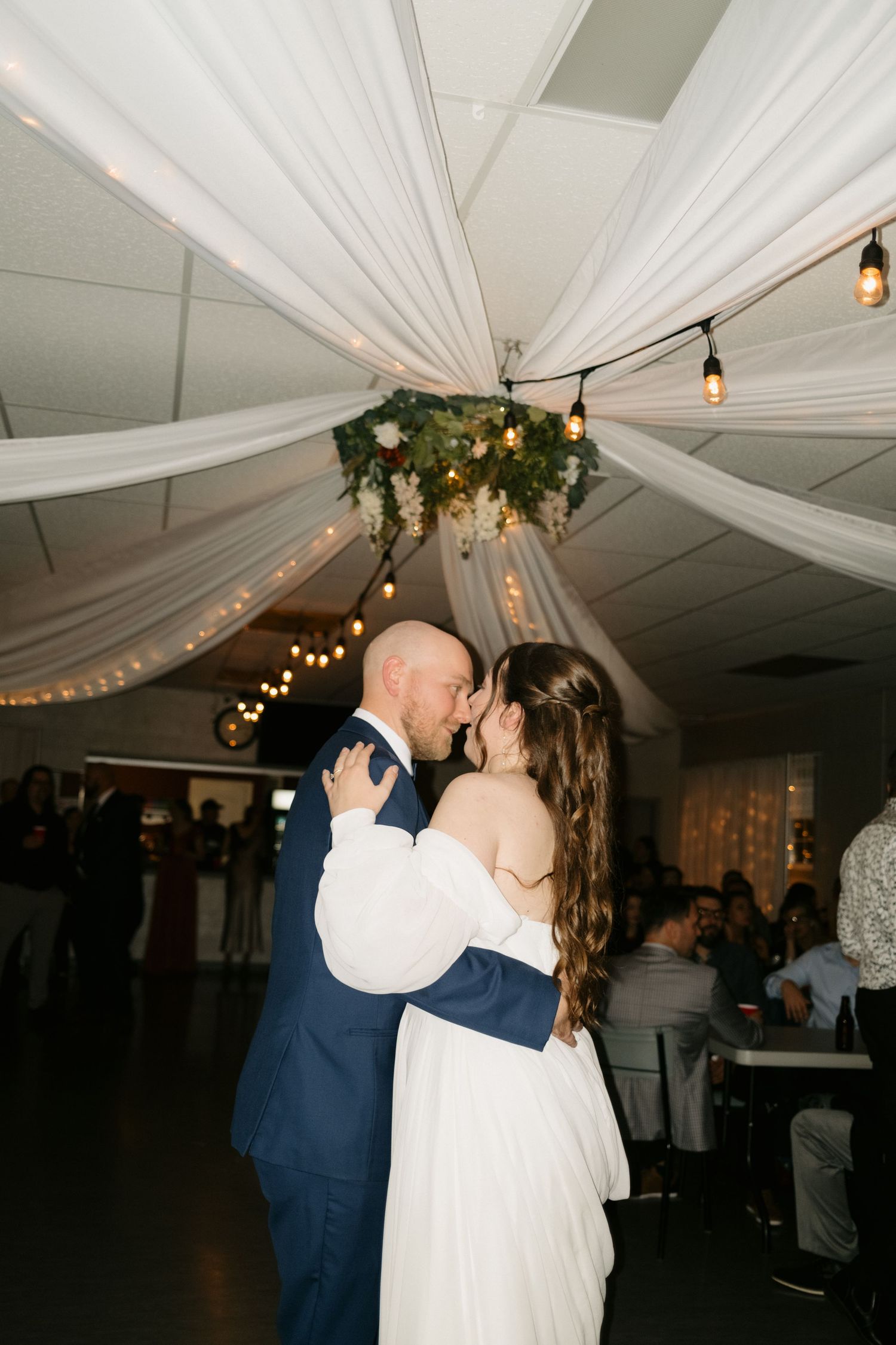 A romantic first dance moment captured at an indoor wedding reception with draped white fabric and string lights overhead.