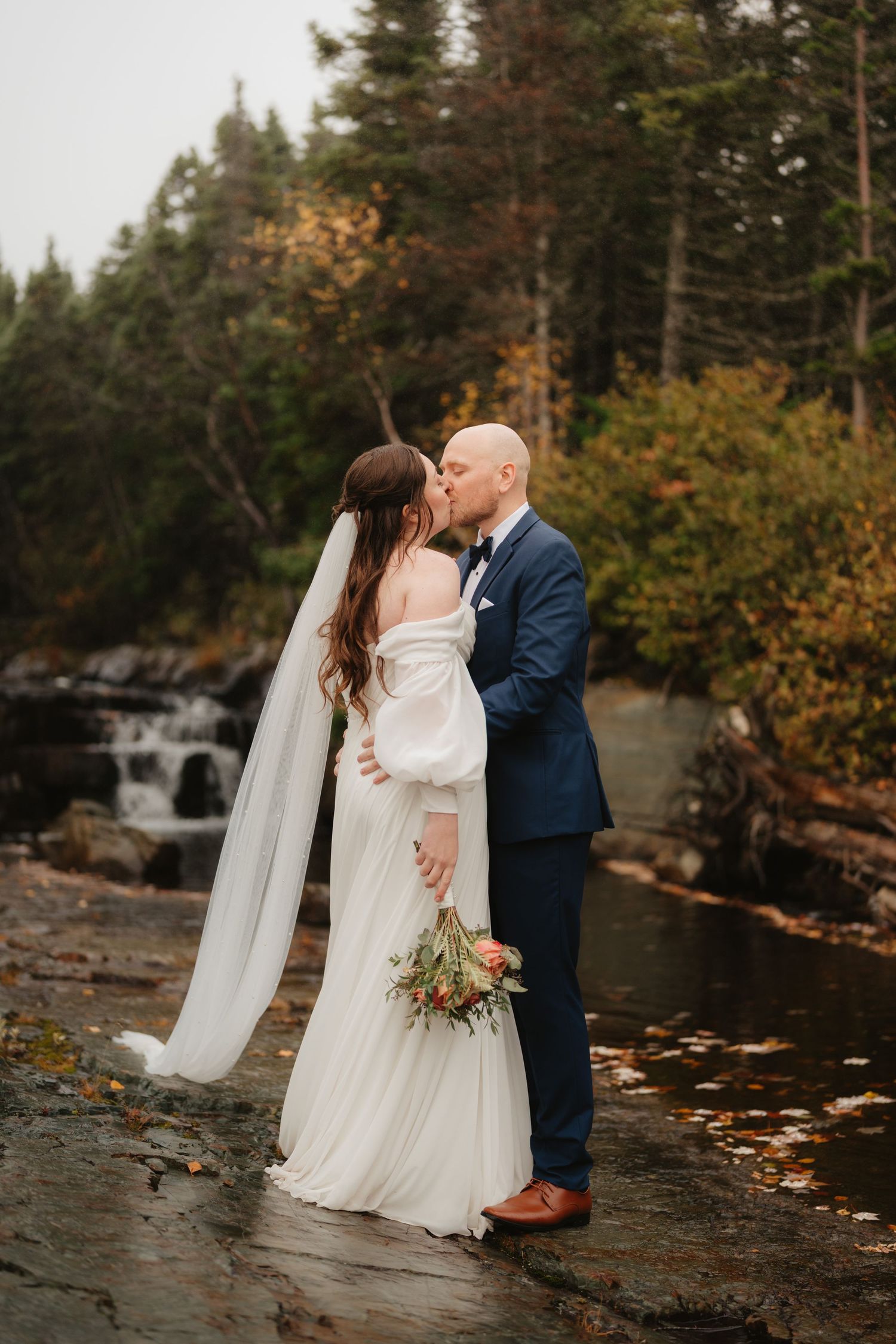 Newlyweds share romantic moments by a scenic forest waterfall during autumn with fallen leaves on rocky shore.