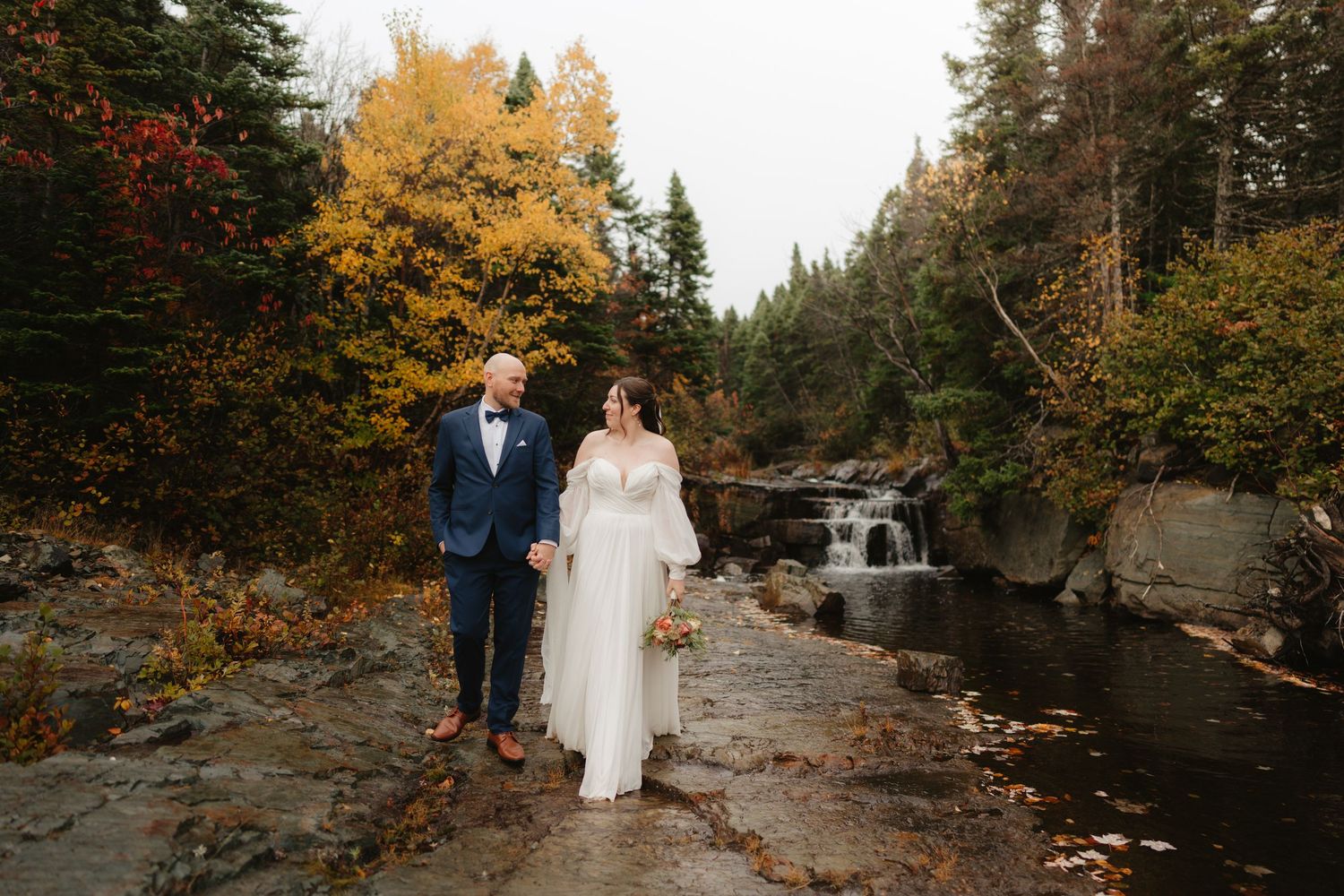 Couple stands together surrounded by vibrant autumn foliage near a rocky riverbank during golden hour.