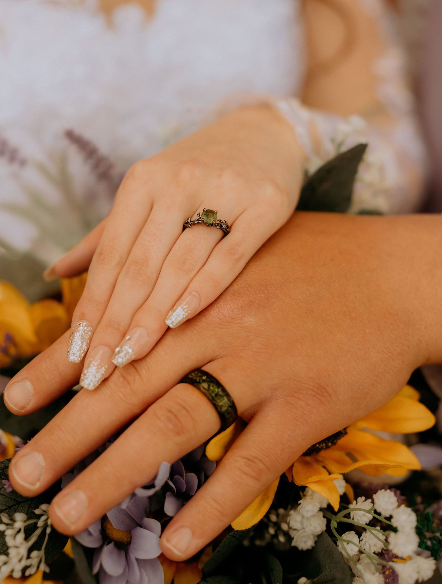 Close-up of manicured hands with engagement ring and band resting on floral arrangement with sunflowers and purple blooms.