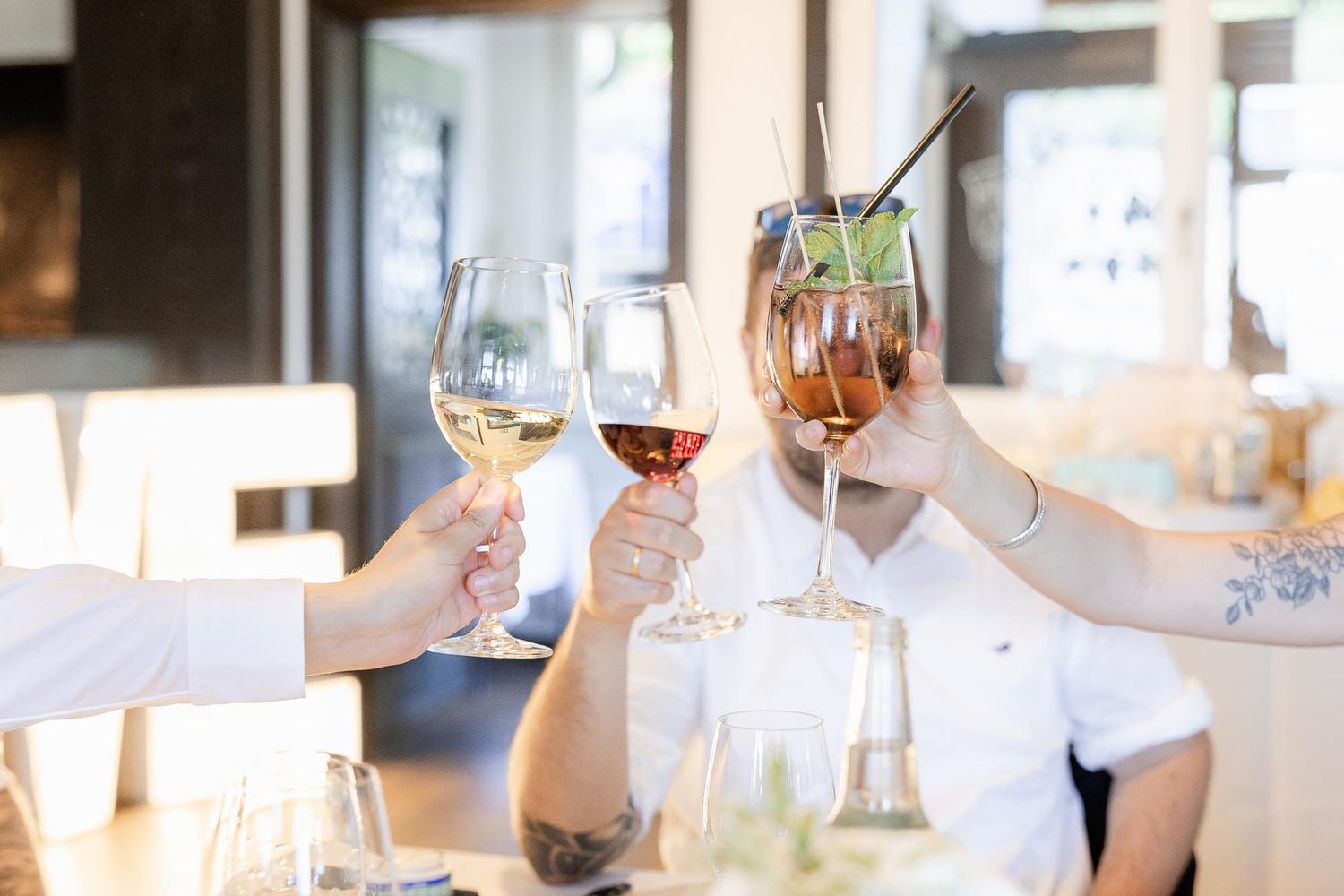 People toasting with wine glasses and a cocktail at a restaurant table in a bright, modern dining setting.