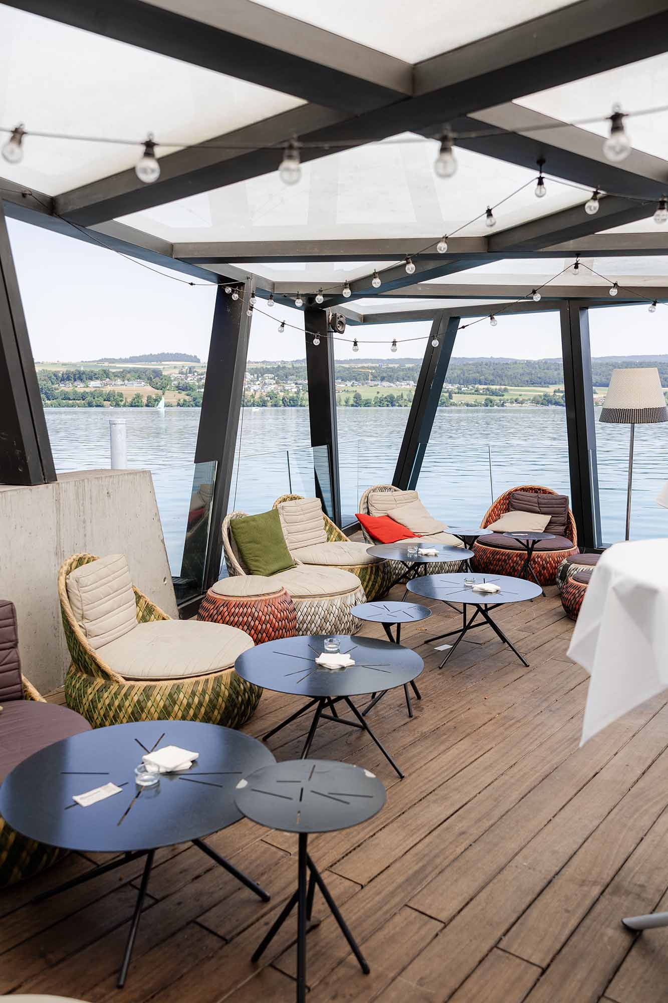 Cozy outdoor lounge area on a boat deck with wicker chairs, small tables, string lights, and scenic river views.