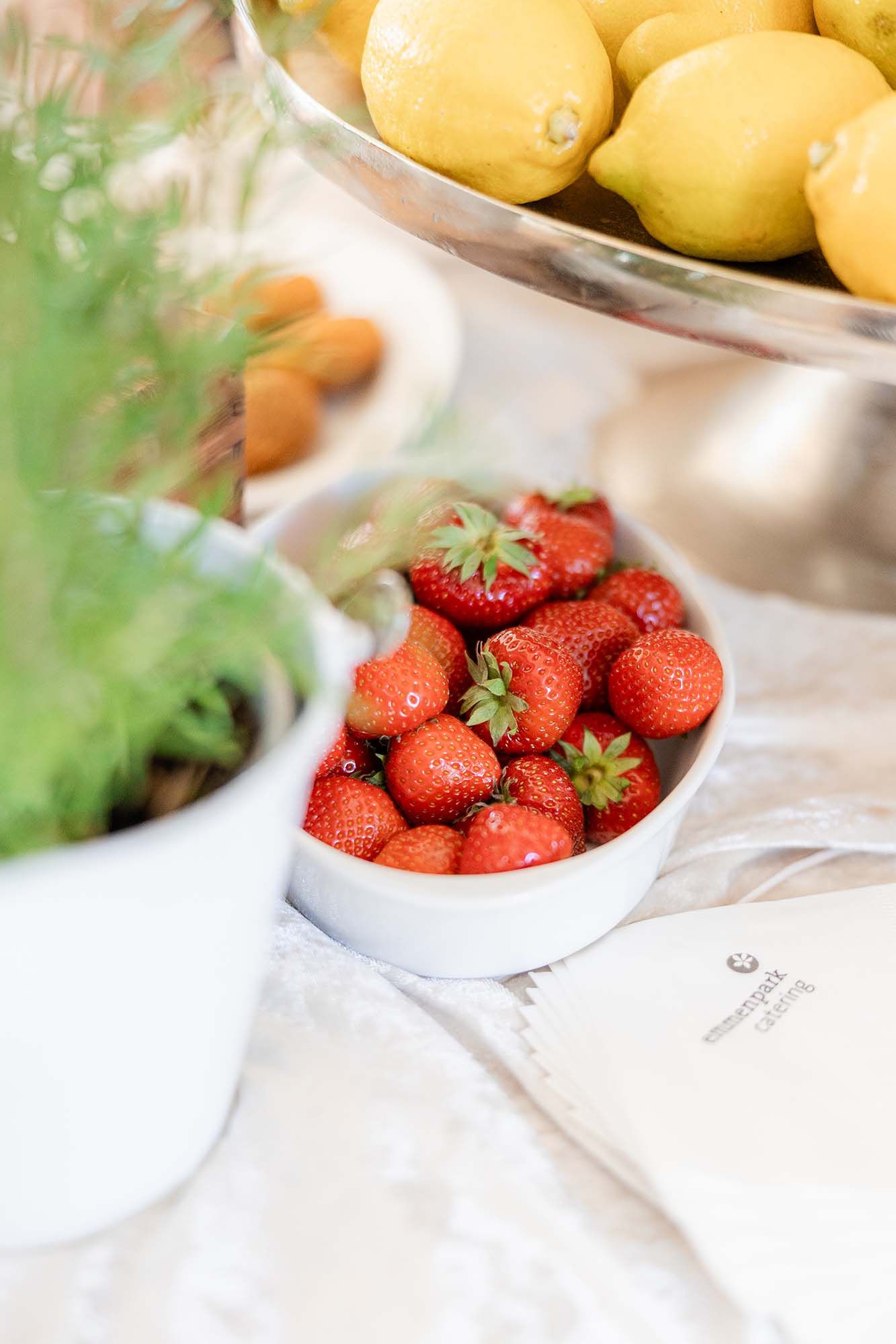 Fresh red strawberries in a white bowl beside lemons in a glass stand and green herbs on a bright table.