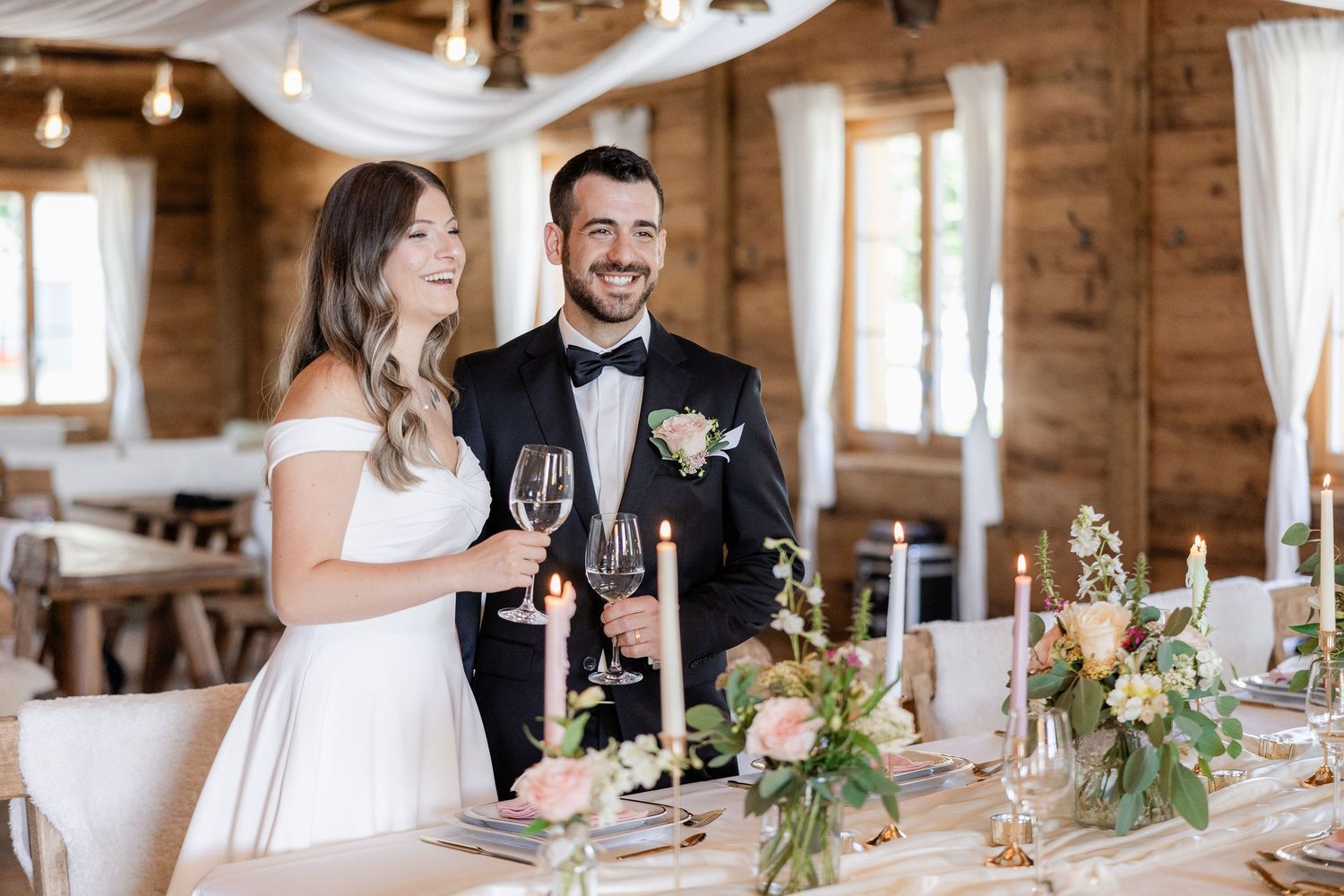 Smiling bride and groom in wedding attire toasting with wine glasses at elegantly decorated reception table.