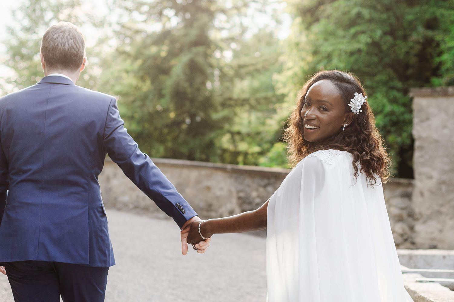 A couple in wedding attire holds hands while walking together through a lush garden setting.