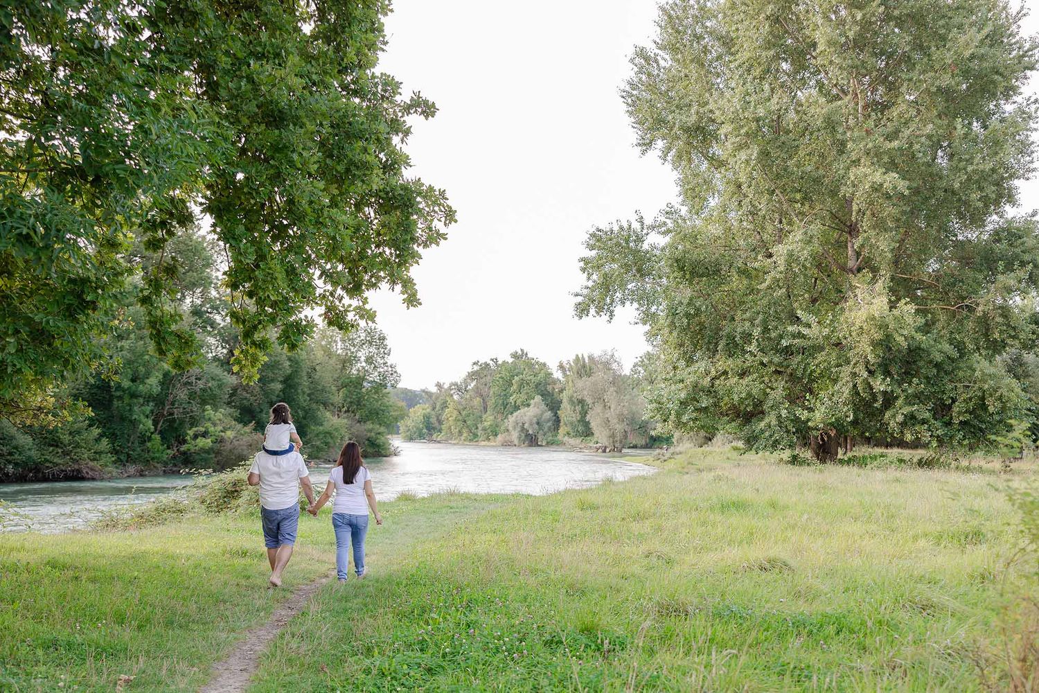 Family walking hand in hand along a scenic river path surrounded by lush green trees and meadow.