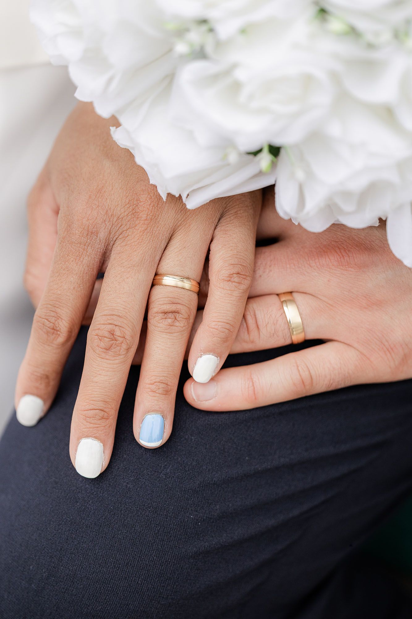 Two hands displaying matching gold wedding bands, with white and blue nail polish, resting near a white floral bouquet.