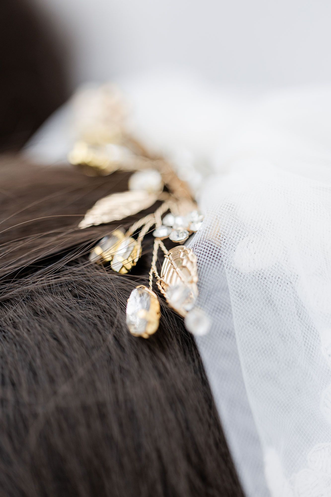 Gold leaf and crystal bridal hair vine with pearl accents nestled in dark hair beside a white wedding veil.