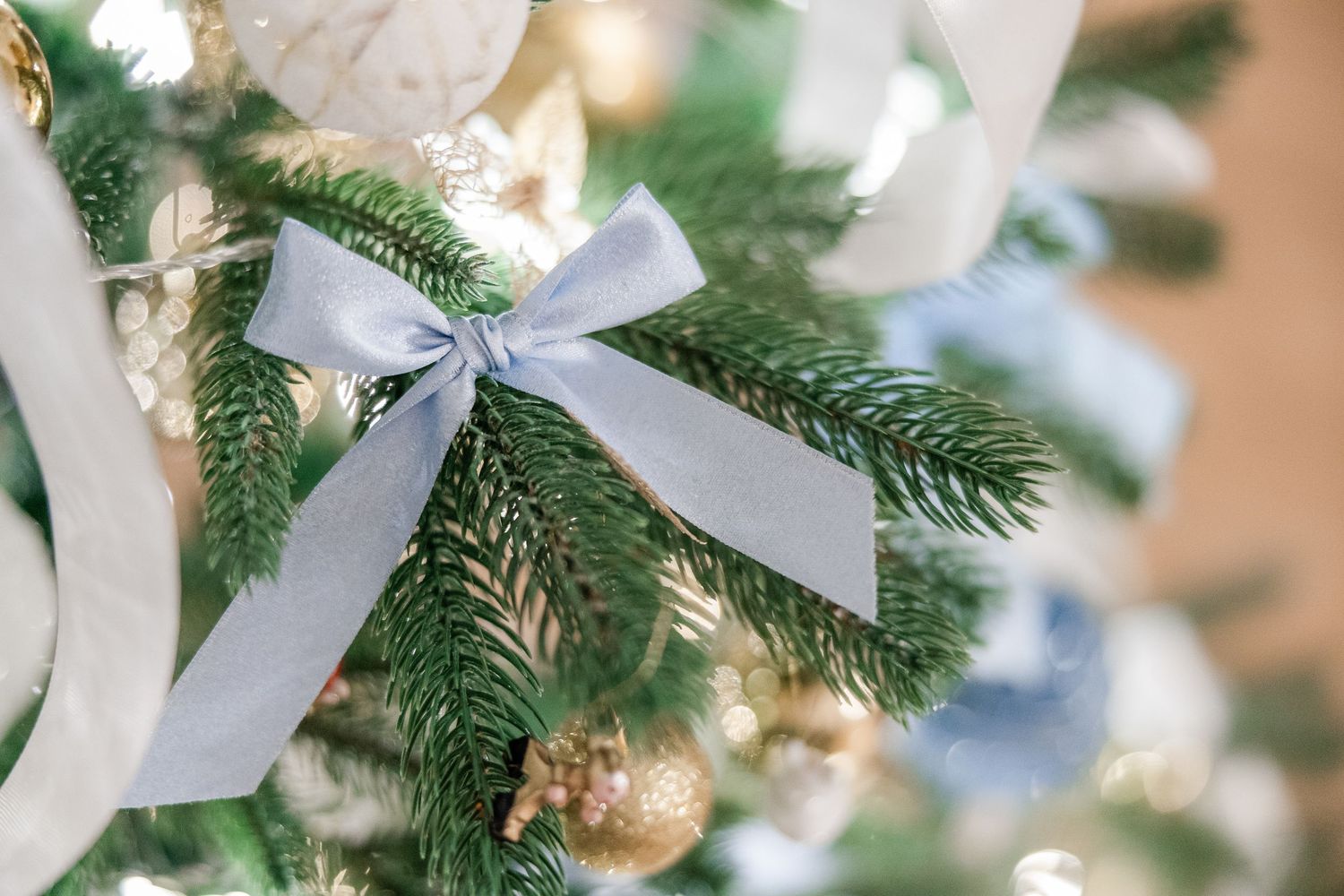 White satin ribbons and gold ornaments decorate a fresh green Christmas tree with warm lighting in the background.