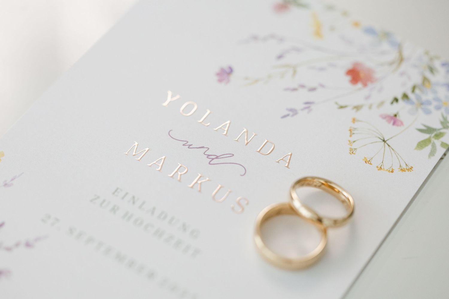 Wedding invitation cards with delicate floral designs displayed alongside gold wedding rings on a white surface.