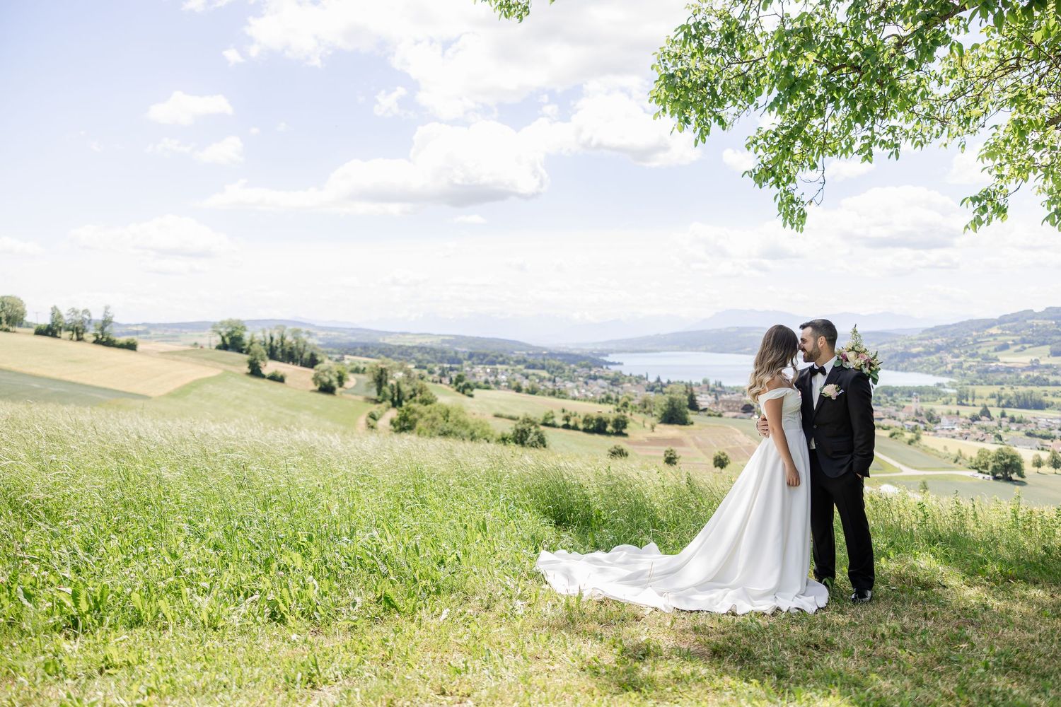 Bride and groom share a romantic moment on a lush green hillside with a stunning lake and valley view.