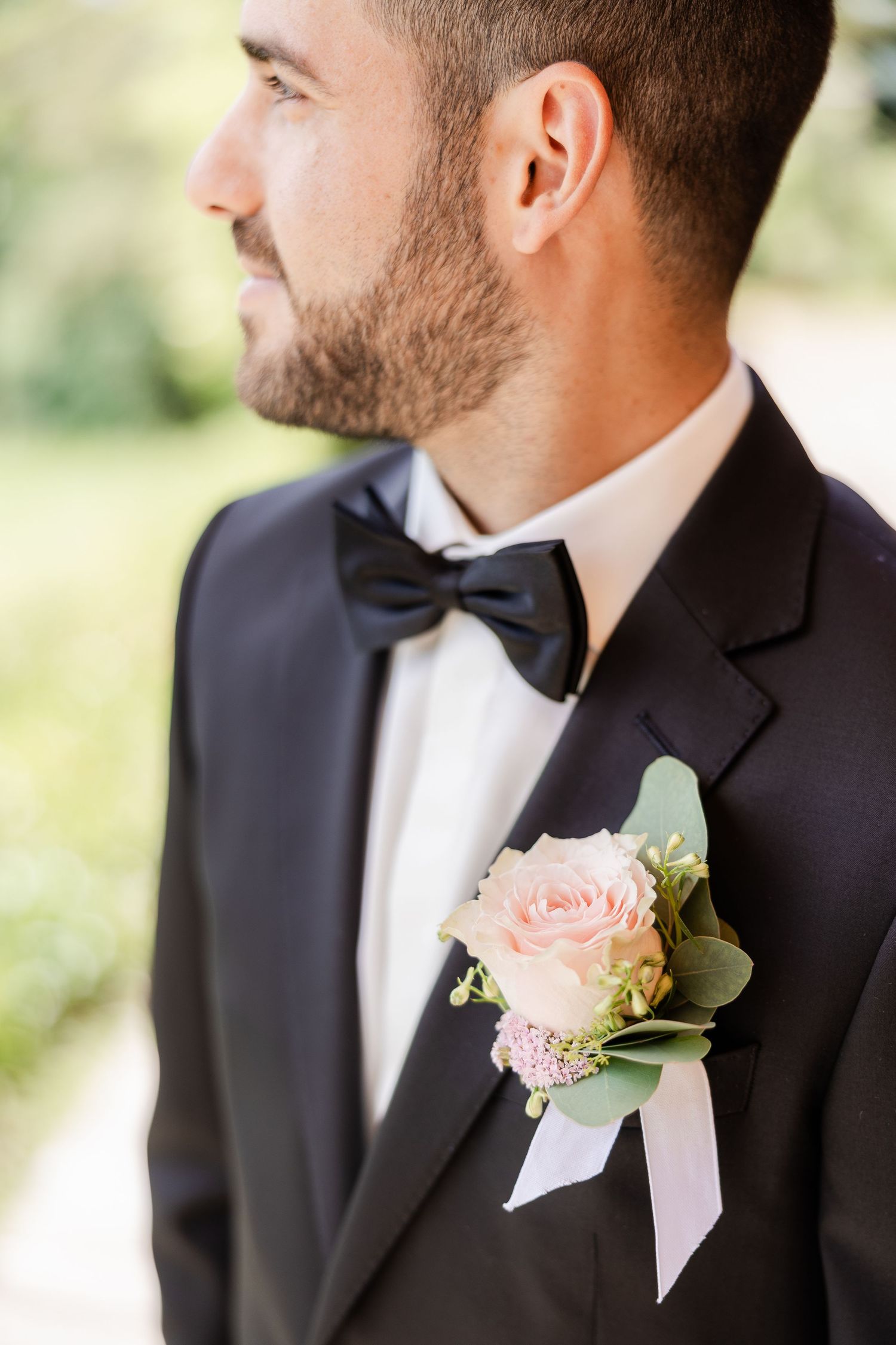 Groom in black tuxedo with bow tie wearing a pink rose boutonniere with greenery and ribbon at a wedding.