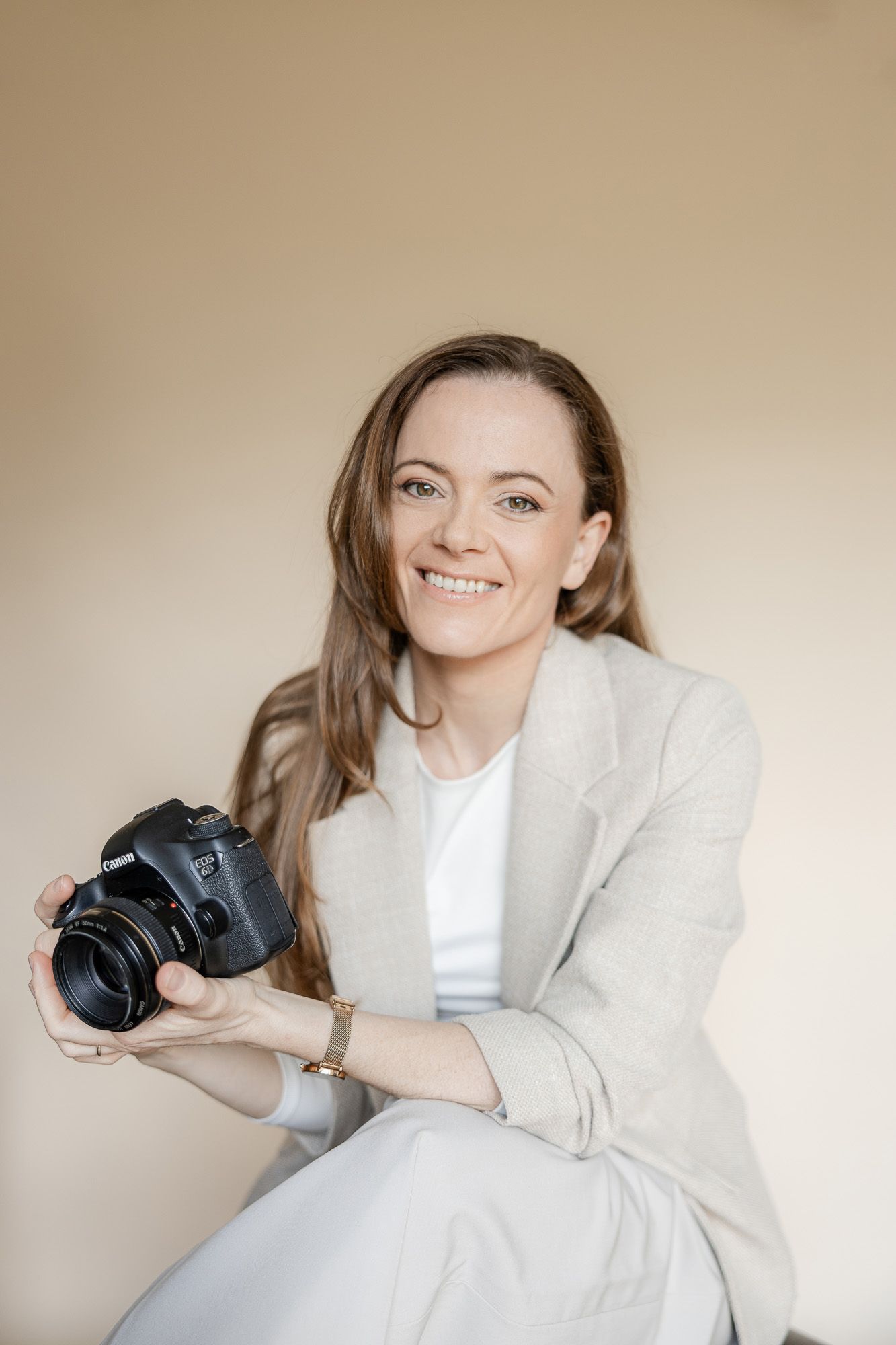A photographer in a white blazer and t-shirt poses playfully with a professional camera against a neutral background.