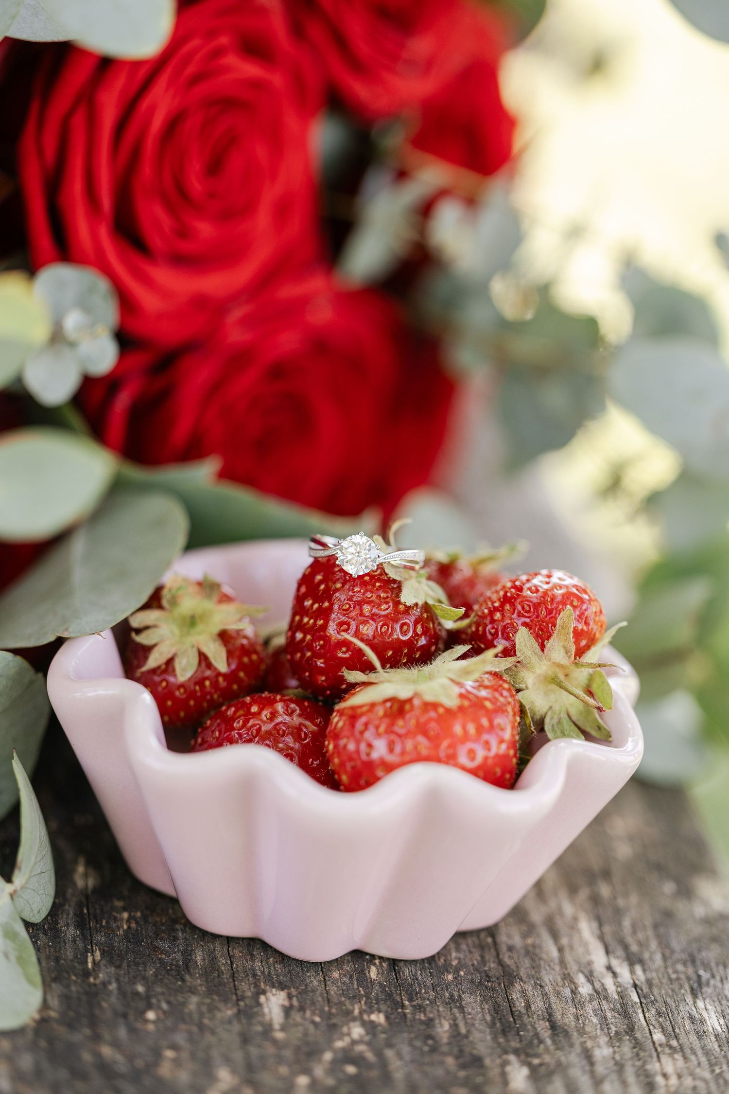 Fresh red strawberries in a pink scalloped bowl with vibrant red roses in the background on rustic wood.