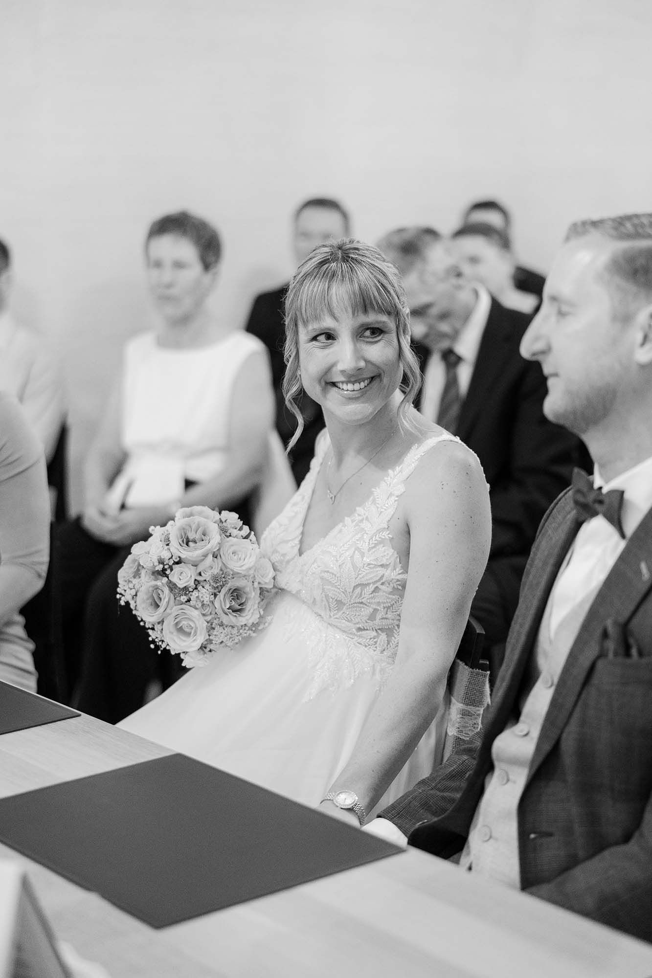 Smiling bride holding a rose bouquet sits beside groom in bow tie during a black and white wedding ceremony.