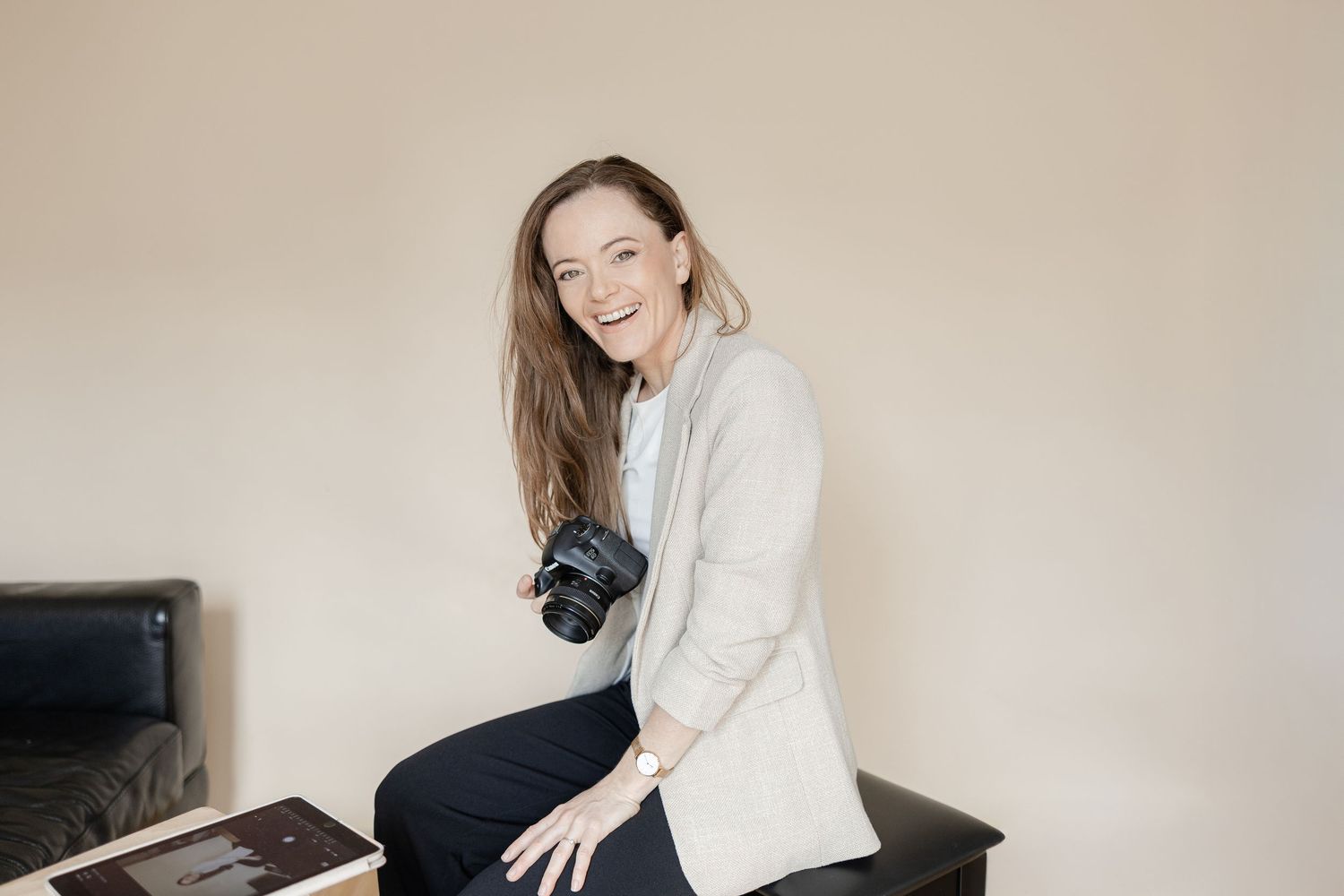 A photographer wearing a grey sweater and jeans sits casually with a camera against a neutral backdrop.