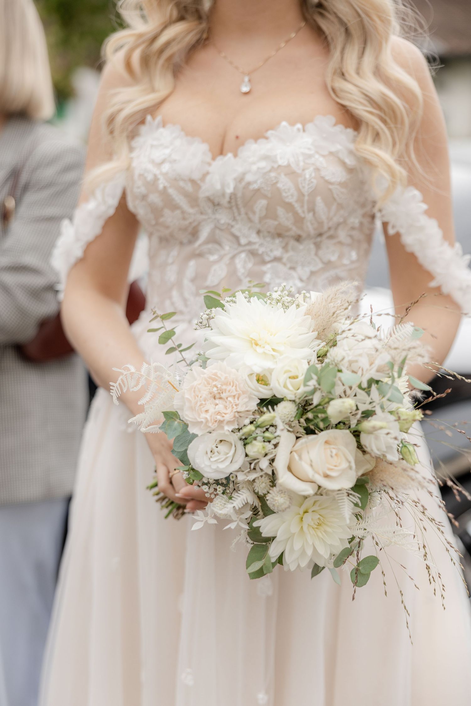 Close-up of a bride in a delicate lace wedding dress holding an elegant white and blush floral bouquet with roses and dahlias.