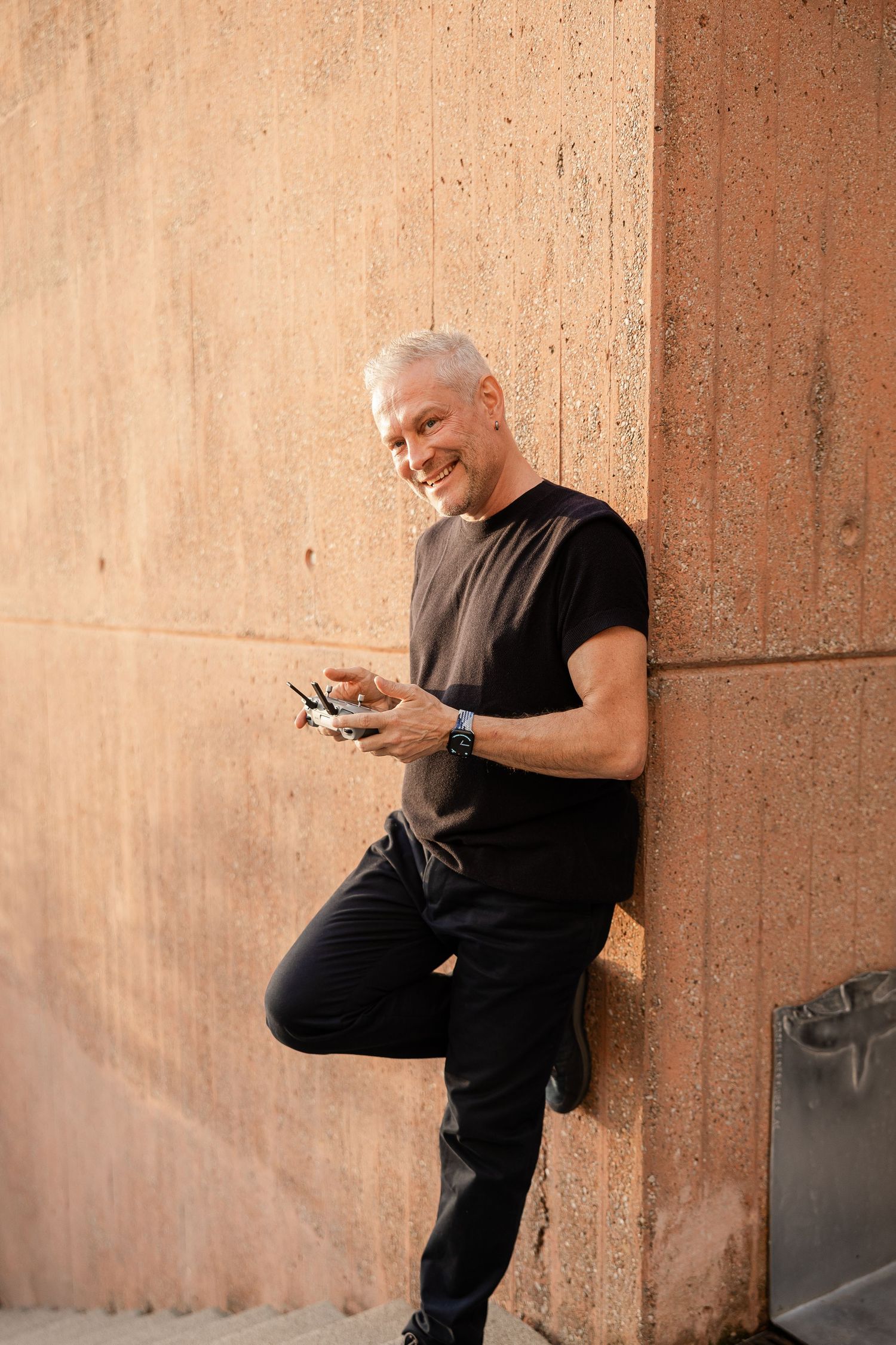 A person in black clothing sits against a textured wall while looking at their phone.